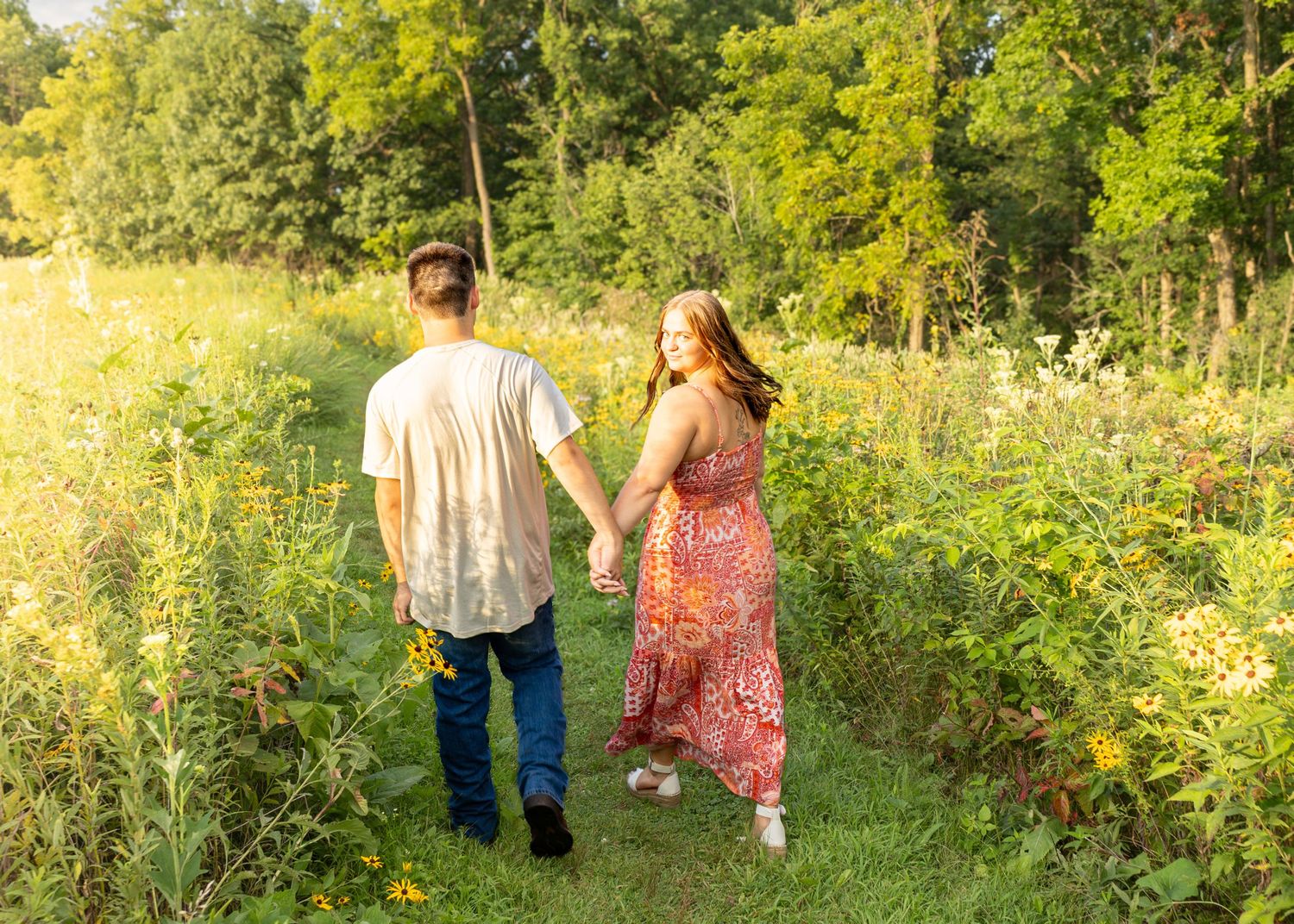 Couple holding hands walking through a sunlit wildflower meadow path surrounded by trees.