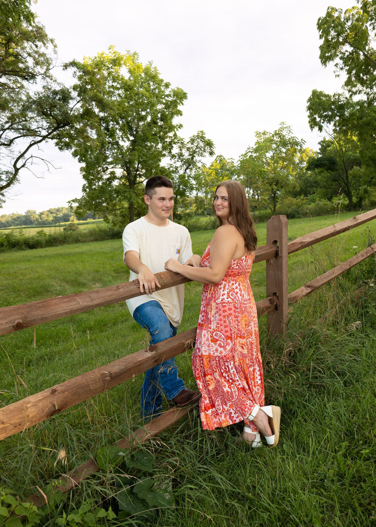 Young couple posing together by wooden fence in lush green field, woman in orange floral dress smiling.