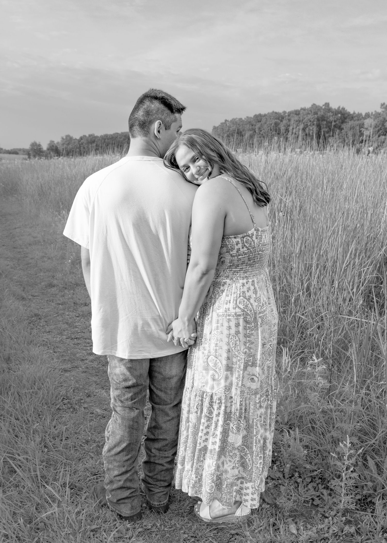 Black and white photo of couple holding hands on field path, woman leaning lovingly on mans shoulder outdoors.