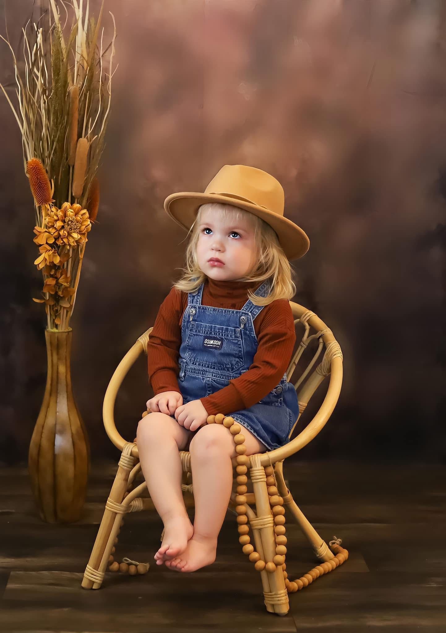 Toddler girl in denim overalls and tan hat sits in rattan chair with autumn backdrop.