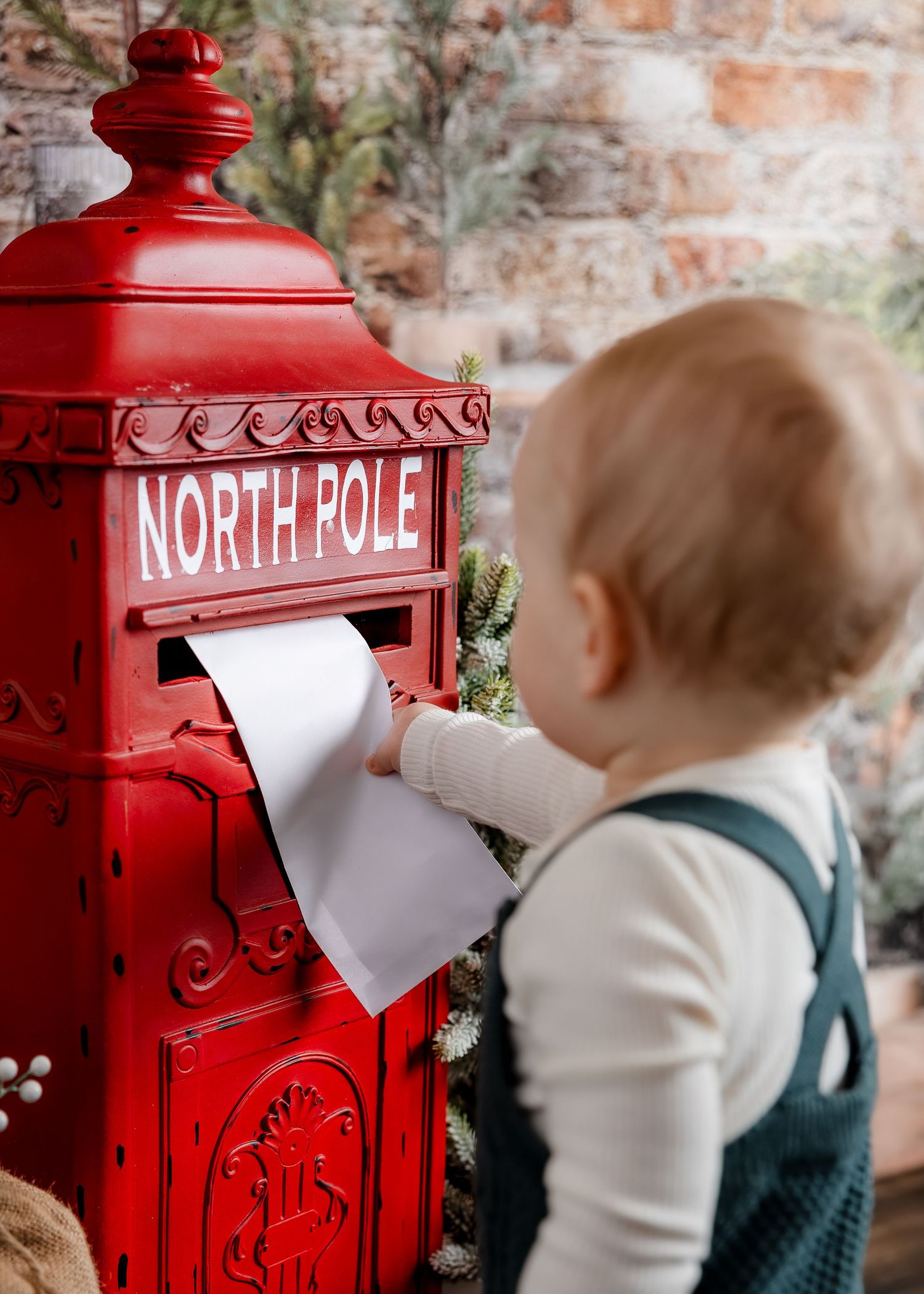 Baby boy in green overalls mailing a letter to Santa in a red North Pole mailbox.