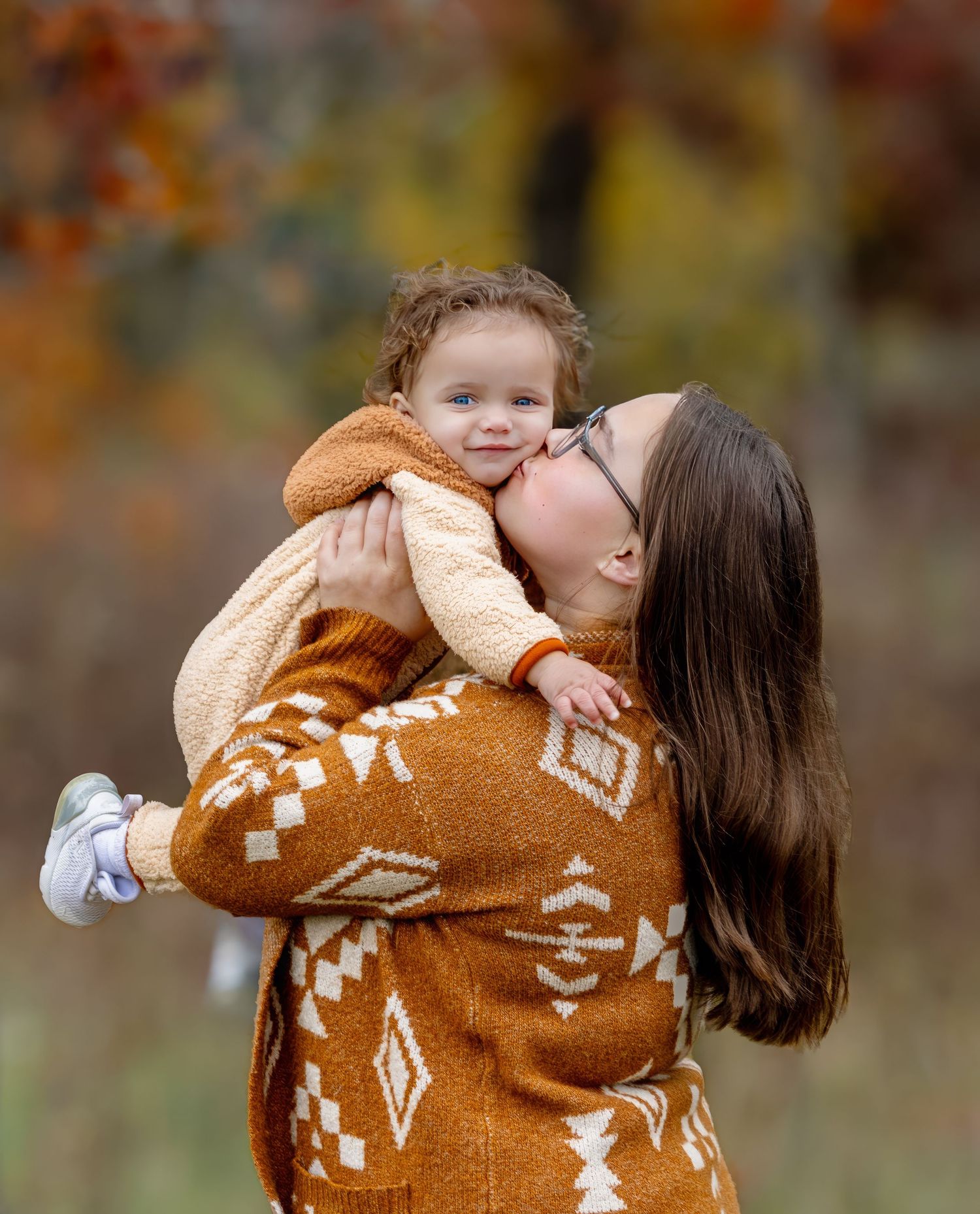 A brunette mother in an orange patterned cardigan kisses her toddler outdoors in fall foliage.