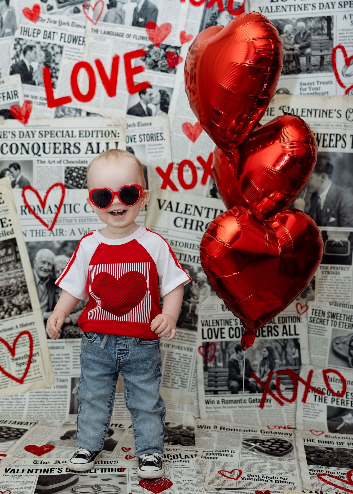 Smiling toddler in red heart shirt stands with red heart balloons on Valentine backdrop.
