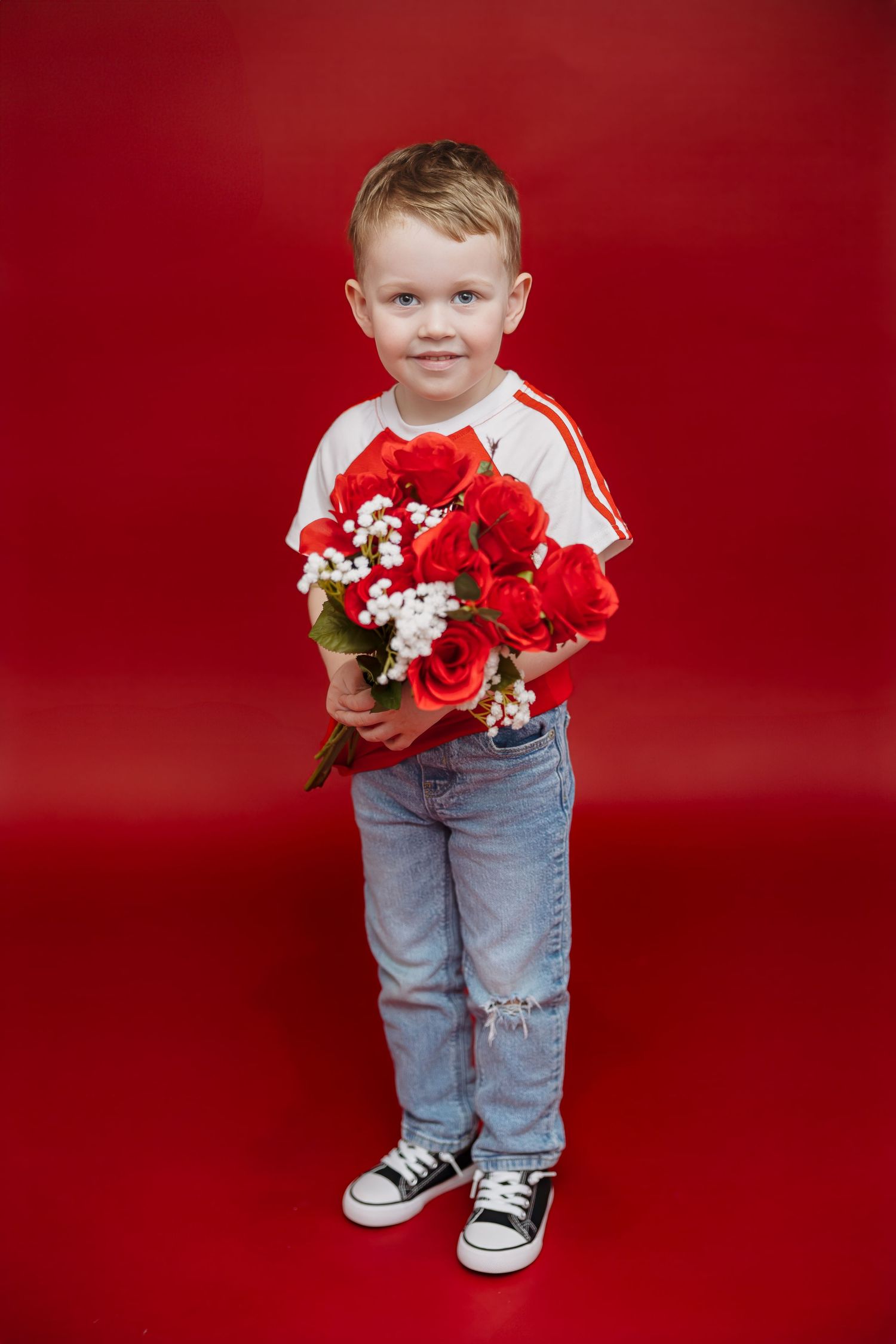 Young boy in red and white shirt holds bouquet of red roses on red background.