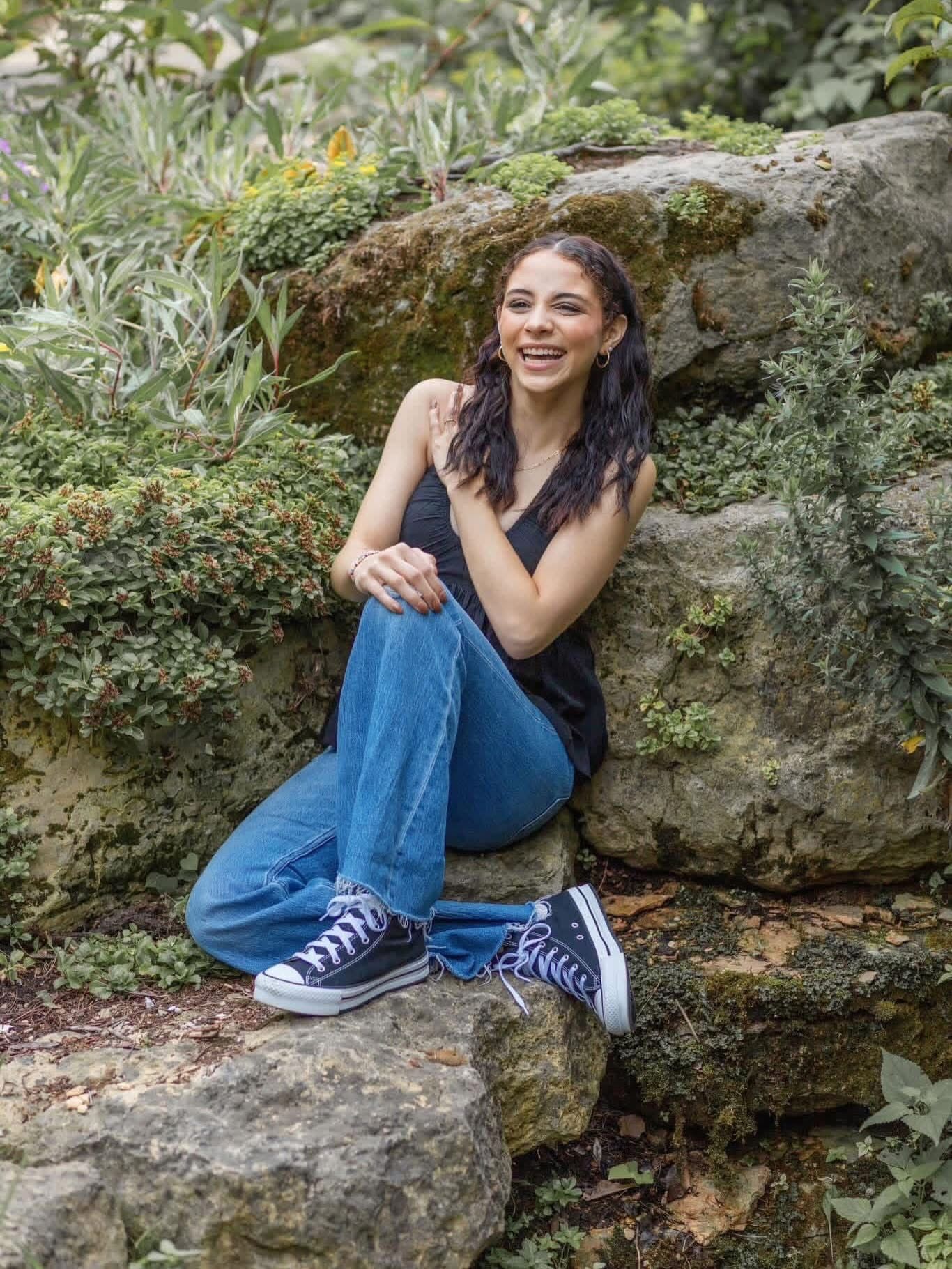 Young woman in jeans and Converse sneakers smiles while sitting on mossy rocks in lush garden.