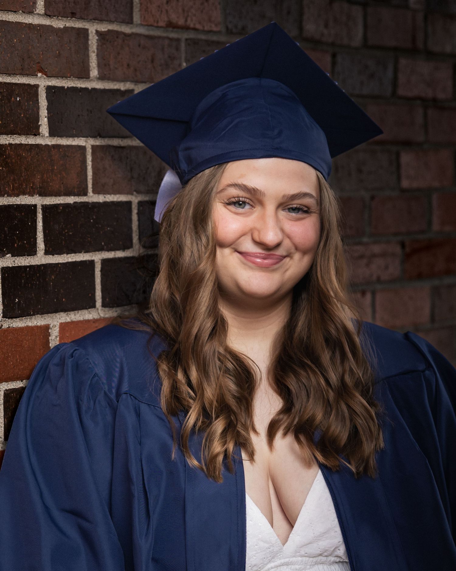 A smiling young woman in a navy blue graduation cap and gown poses against a brick wall.
