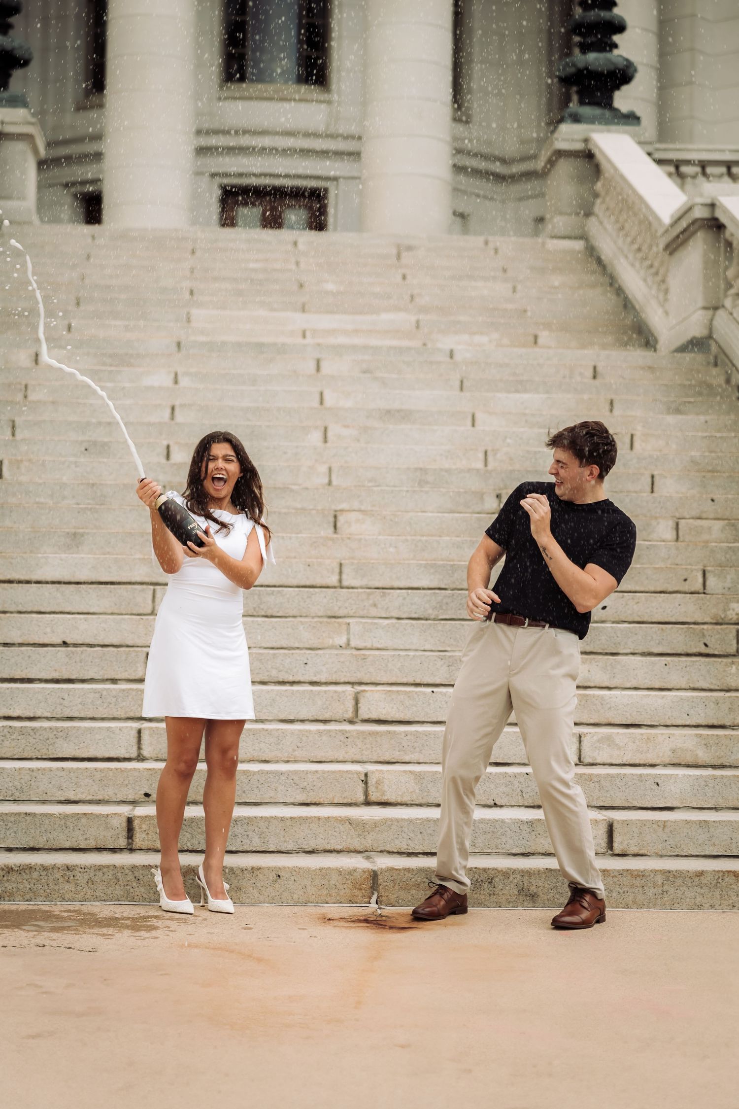 Couple celebrating with champagne on grand steps, woman in white dress spraying bottle while man dances away.