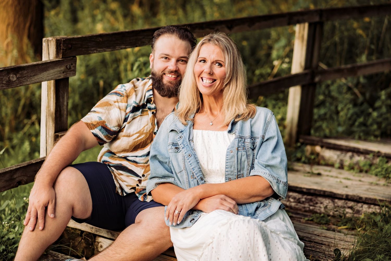 A smiling couple sitting together on a wooden bench outdoors, surrounded by lush green trees.