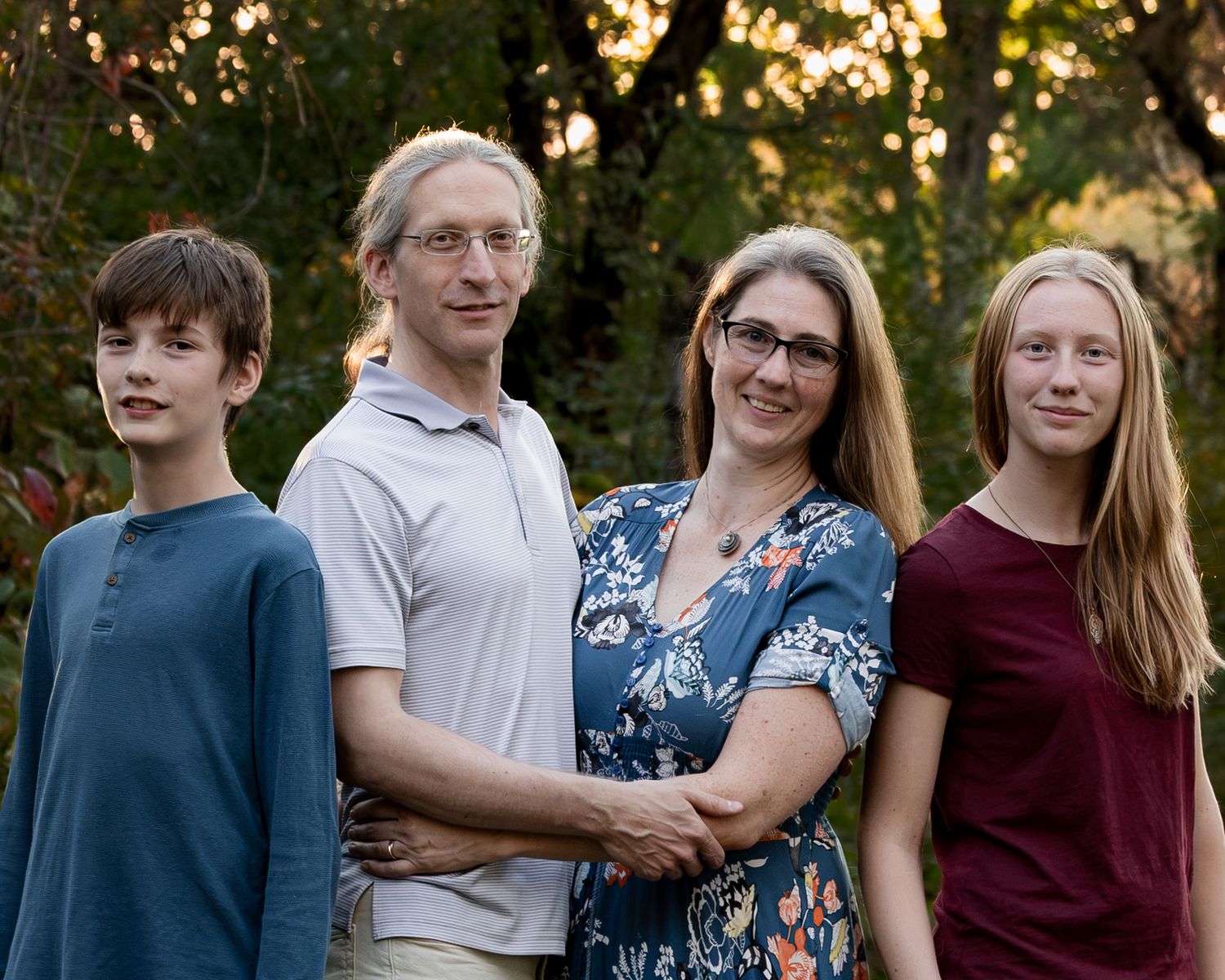A family of four stands close together outdoors among green trees, smiling, with two parents in the middle and two children on either side.