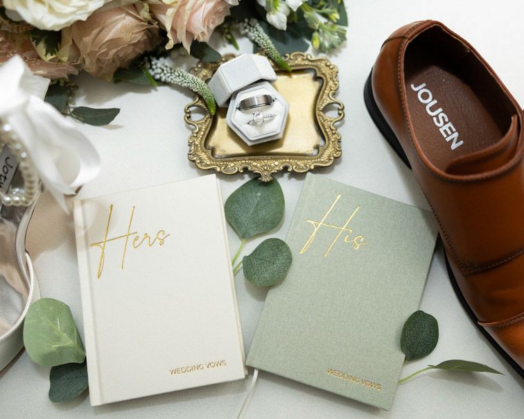 Wedding flat lay with His and Hers vow books, a diamond ring in a gold tray, a brown Jousen leather shoe, and floral and eucalyptus accents.
