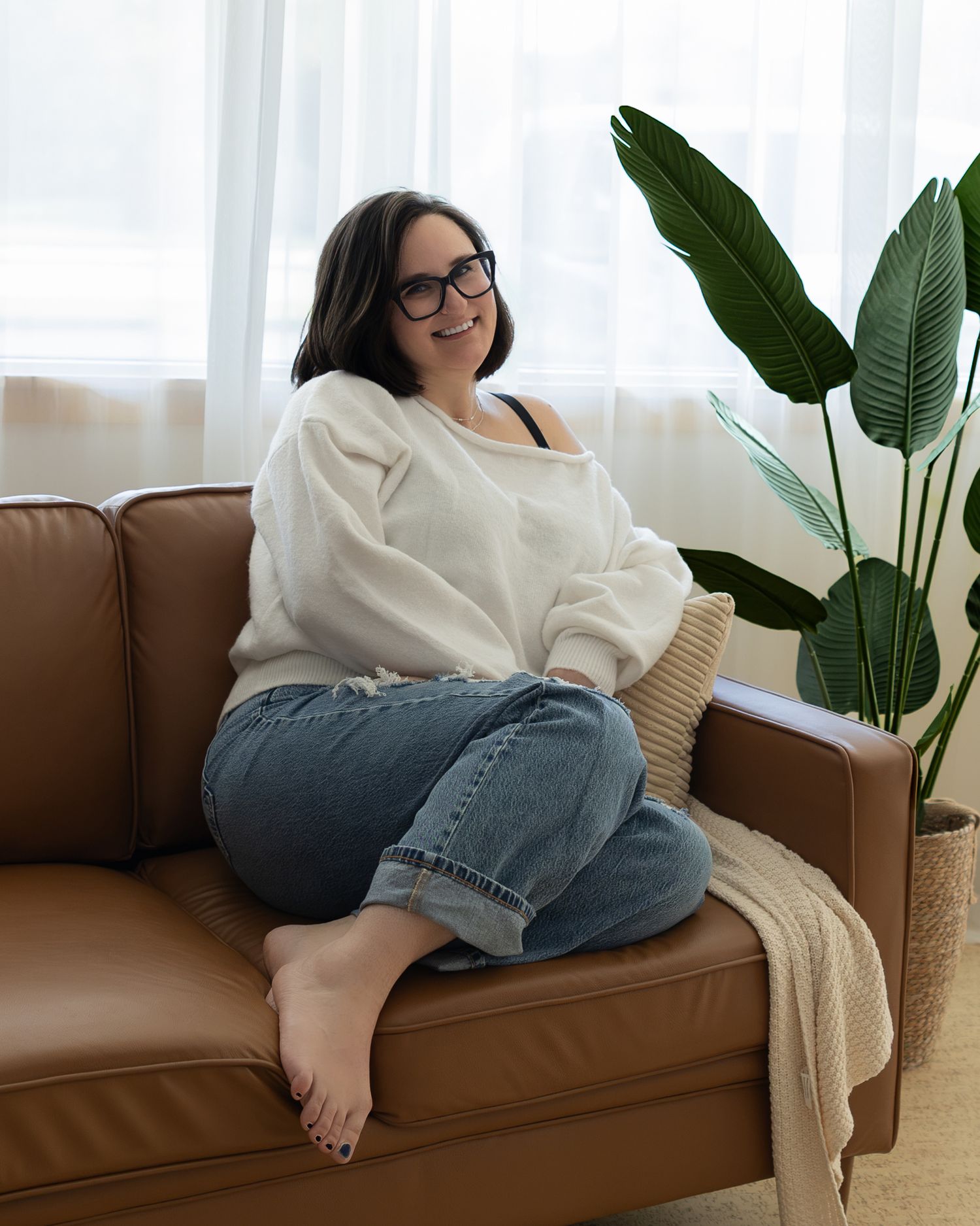 Woman in white sweater and jeans sitting cross-legged on brown leather sofa near large tropical plant.