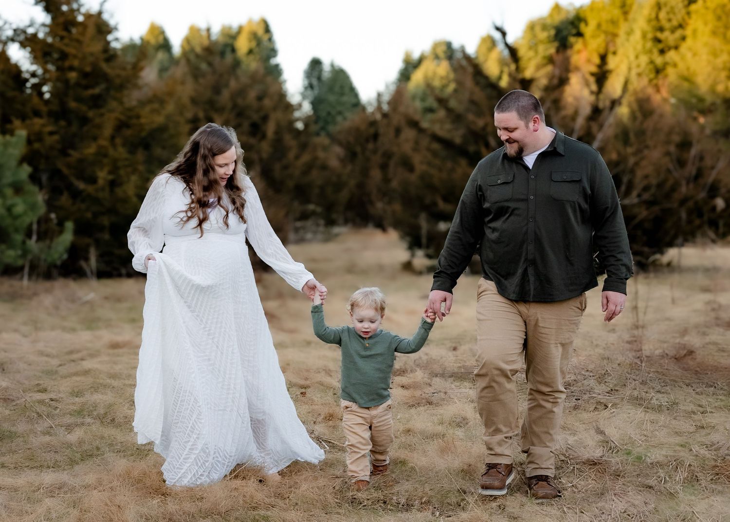 Pregnant couple walking hand-in-hand with toddler son through a dry grass field at sunset.
