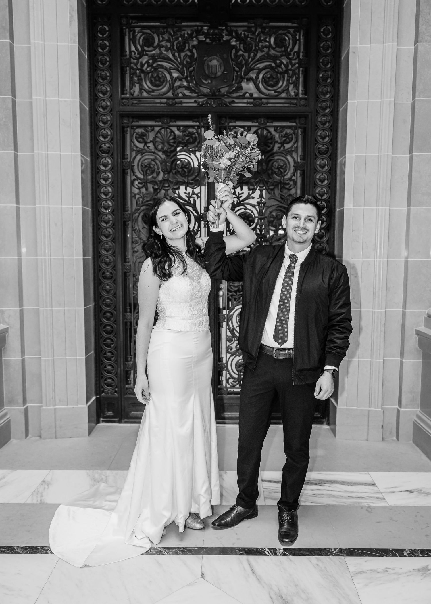 Bride in white gown and groom in black jacket celebrating with raised bouquet at ornate doors.