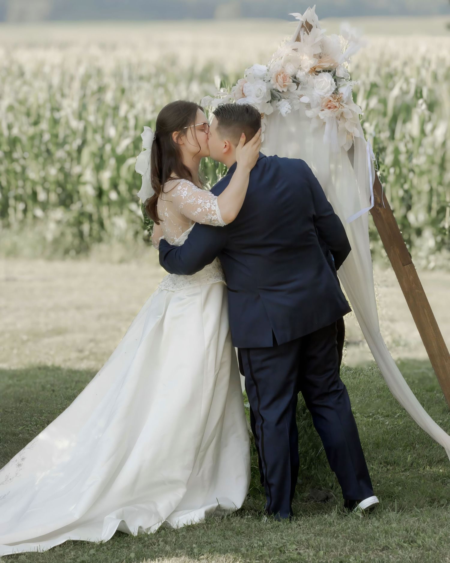 Bride and groom sharing first kiss at outdoor wedding ceremony with floral arch and cornfield backdrop.