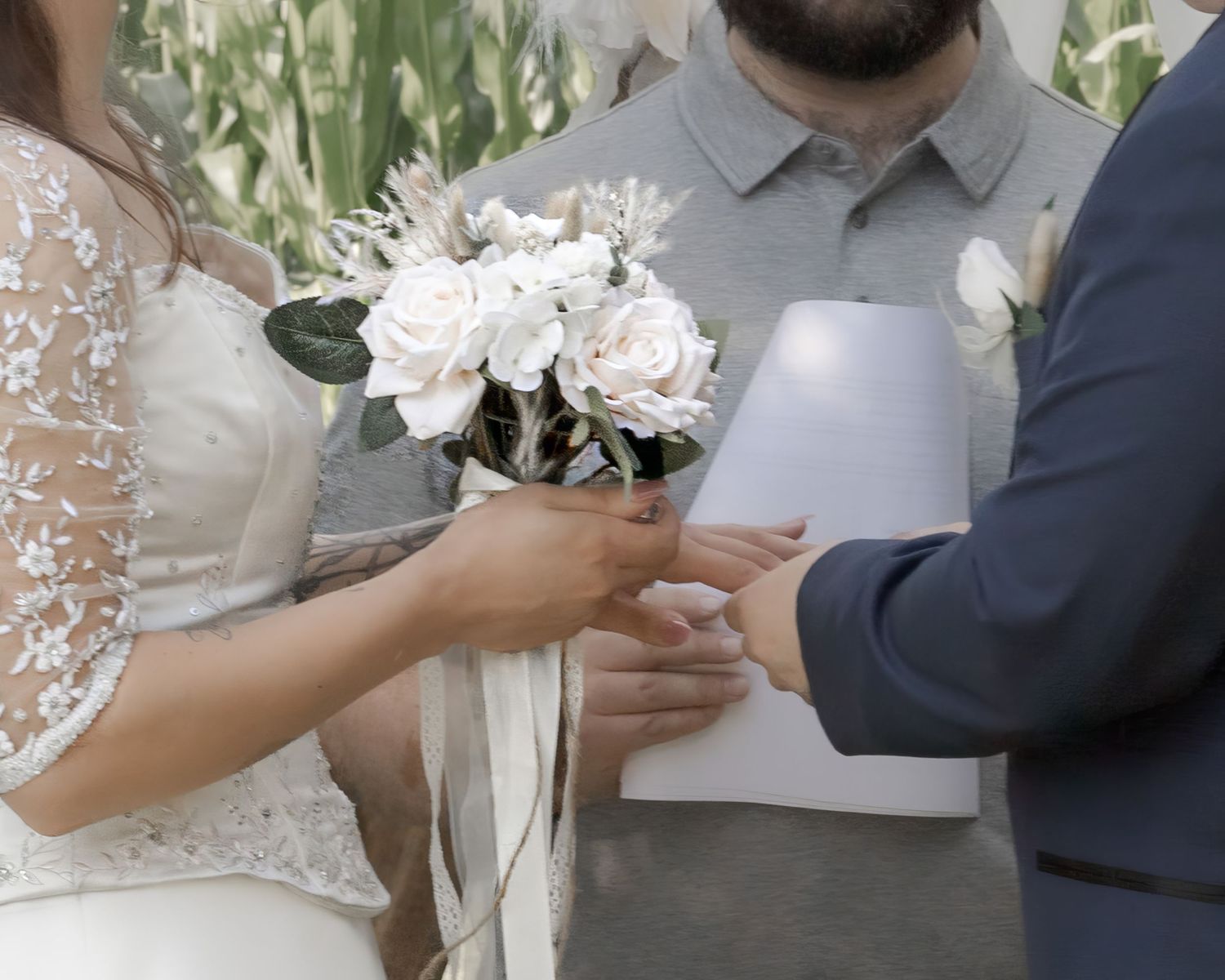 Close-up of bride holding white rose bouquet while groom places wedding ring on her finger during ceremony.