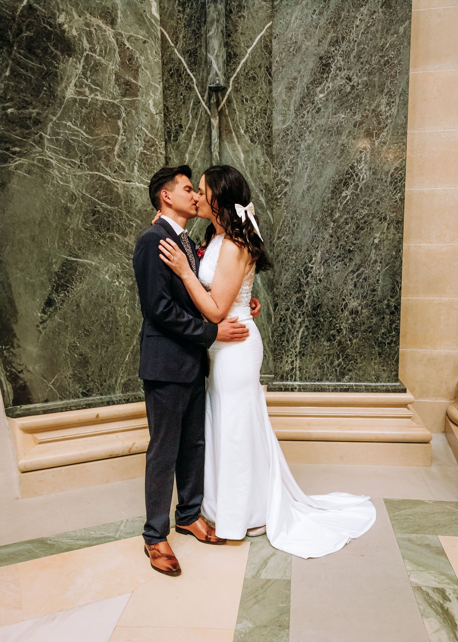 Bride and groom share tender kiss before green marble wall, bride wearing white gown with bow.