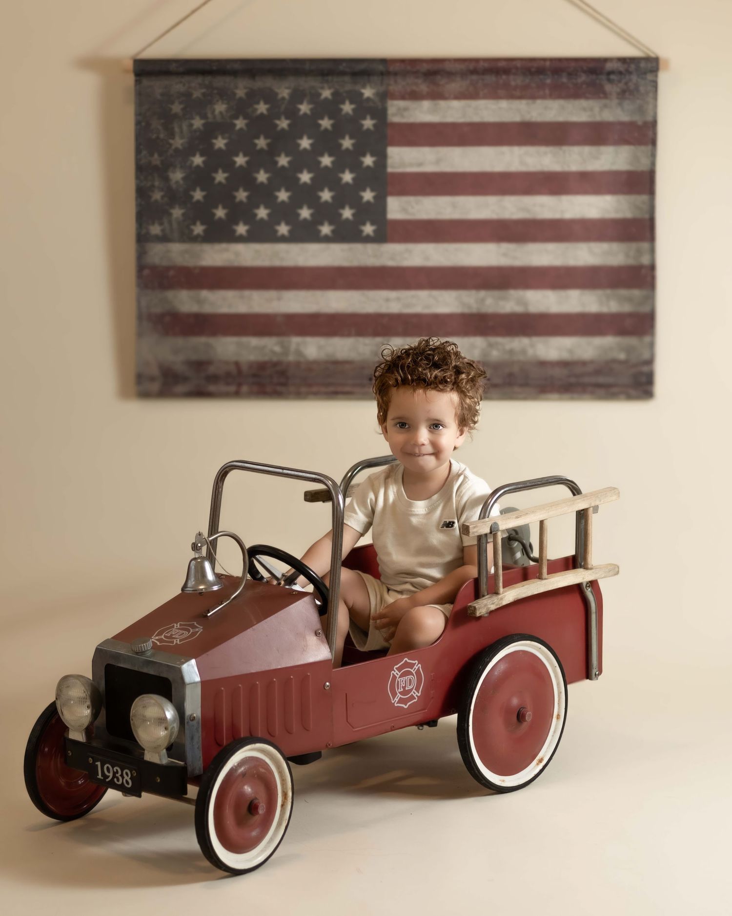 Curly-haired child sitting in a 1938 vintage red pedal fire truck below US flag.