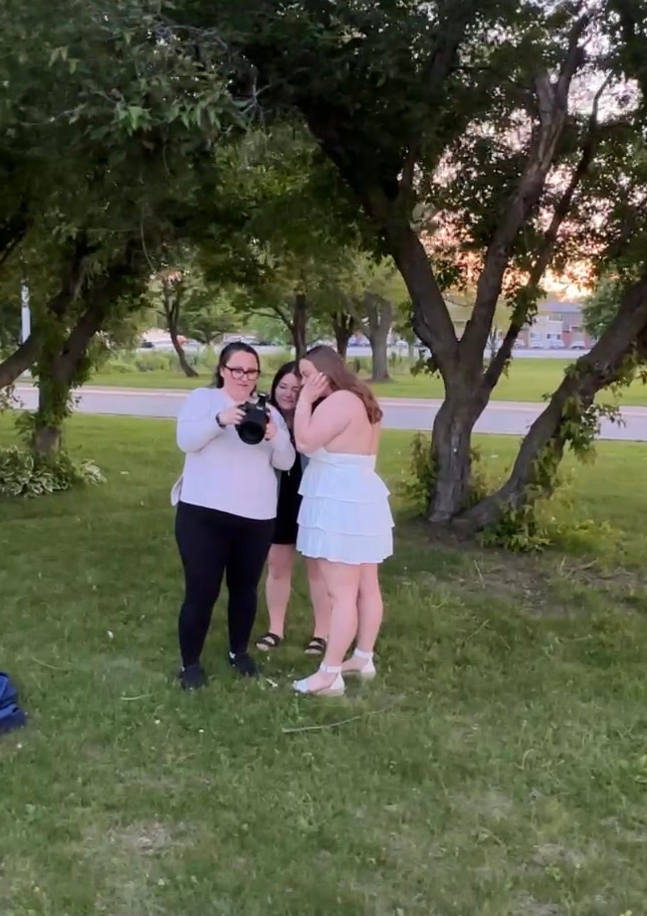 Three women gather outdoors at sunset, viewing photos on a camera near green trees on a grassy lawn.