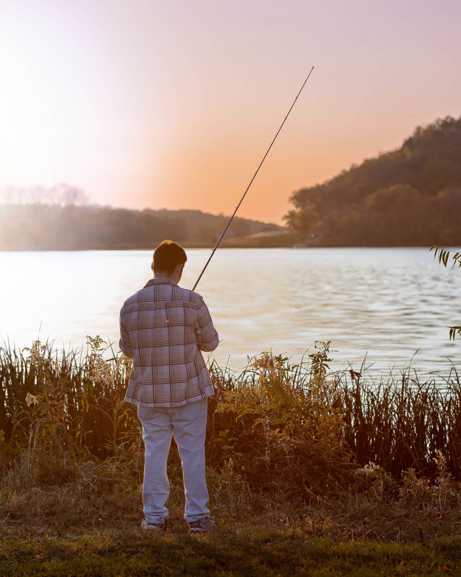 A man fishing at a serene lake during golden hour sunset, standing on grassy shore with fishing rod cast over calm water.