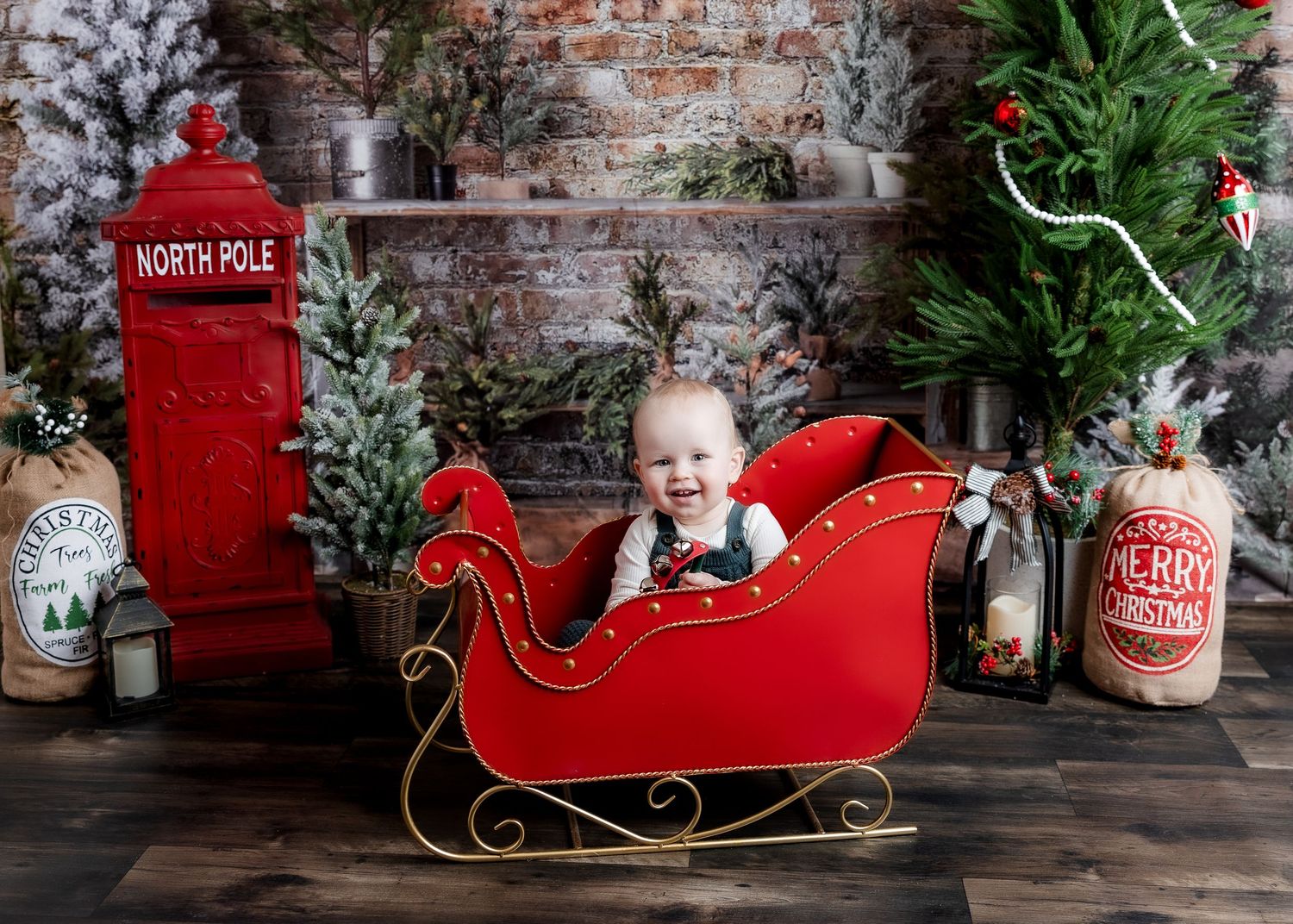 Happy baby boy laughing in red Christmas sleigh surrounded by North Pole holiday decorations.