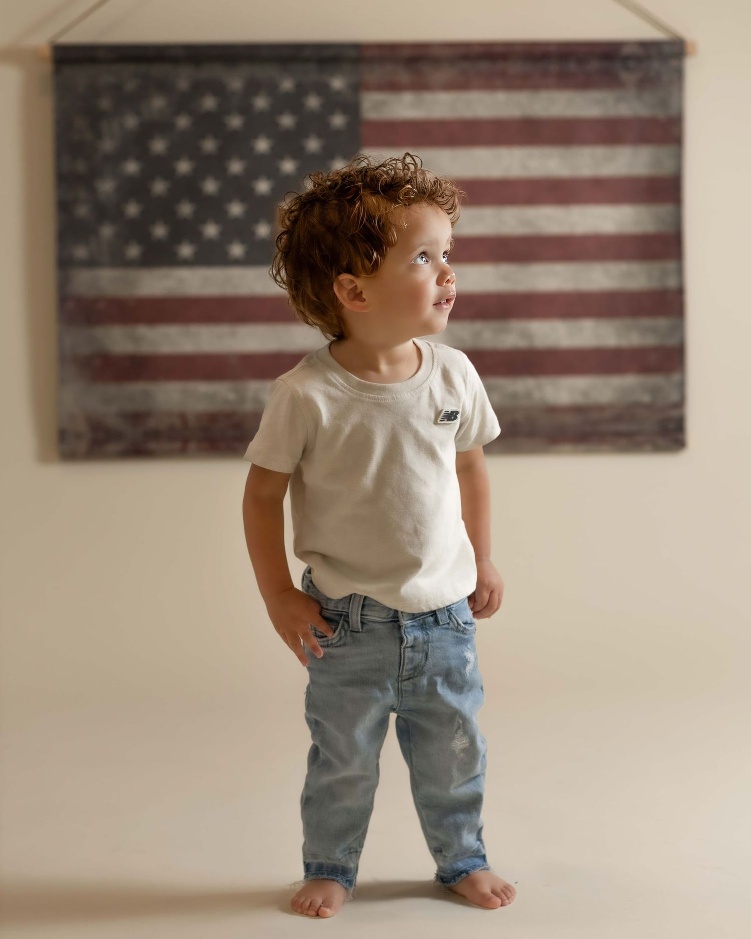 Toddler boy in white tee and jeans stands barefoot before vintage American flag wall art.
