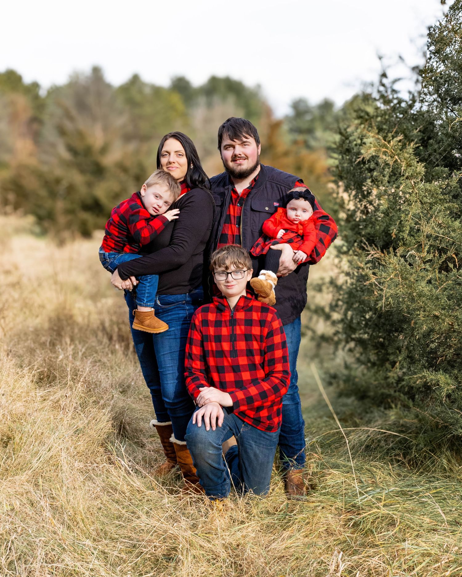 Family of five in red plaid outfits poses among evergreen trees at a tree farm.