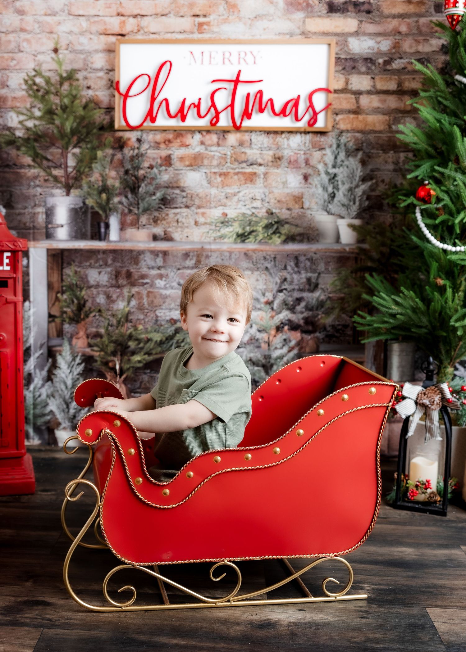 A smiling toddler boy sits in a red and gold sleigh with a Merry Christmas sign and holiday decor behind him.