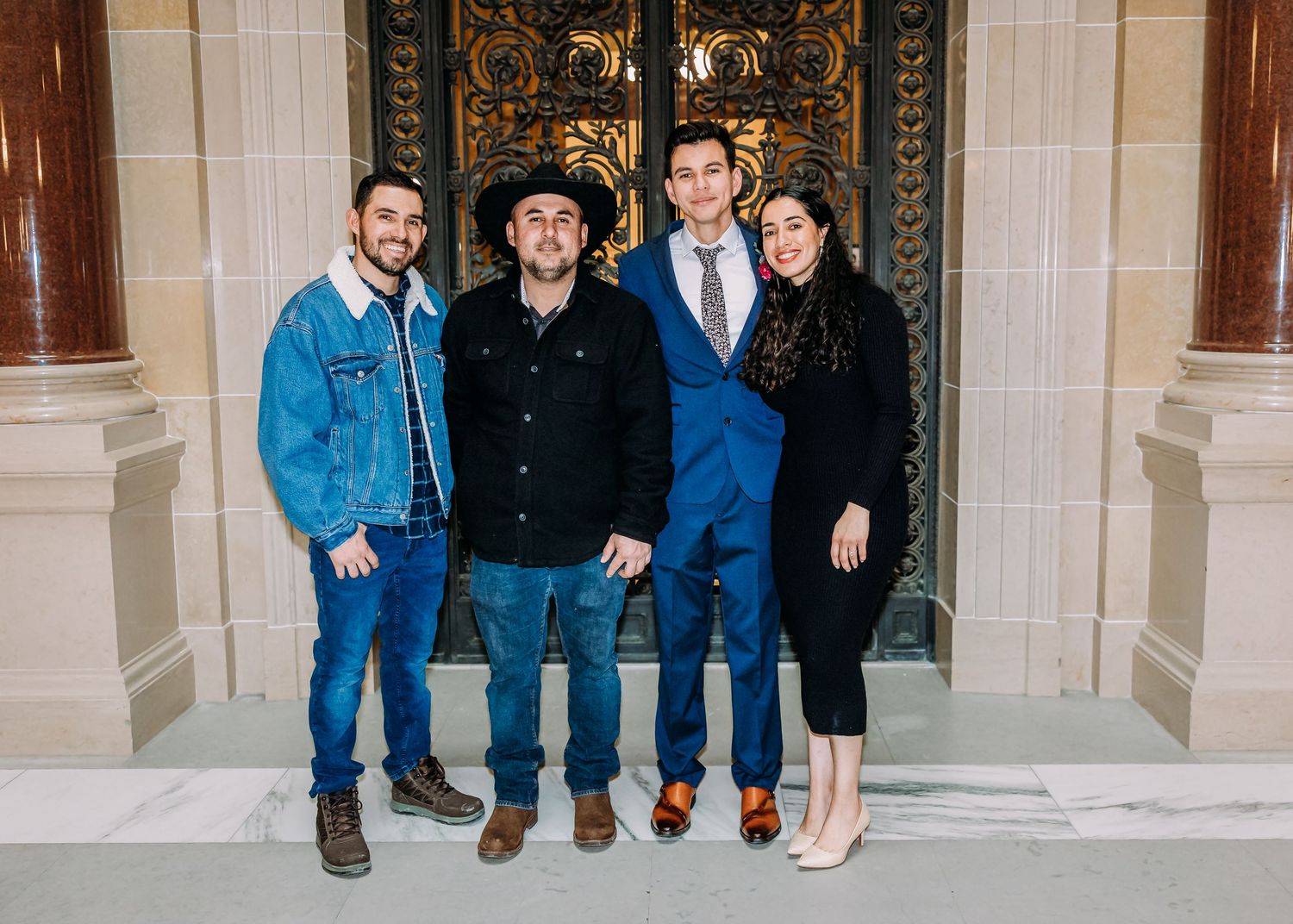 Four guests including two men in casual attire pose before golden ornate doors at a wedding celebration.