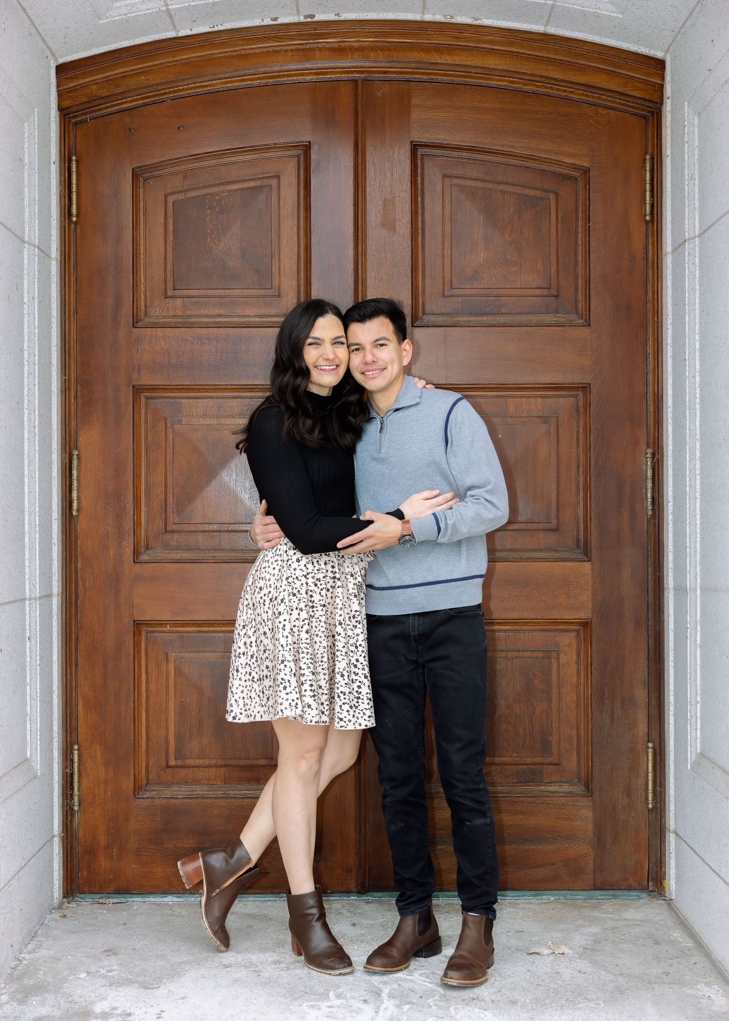 Couple poses happily in front of large wooden double doors, woman in floral skirt and man in gray sweater.