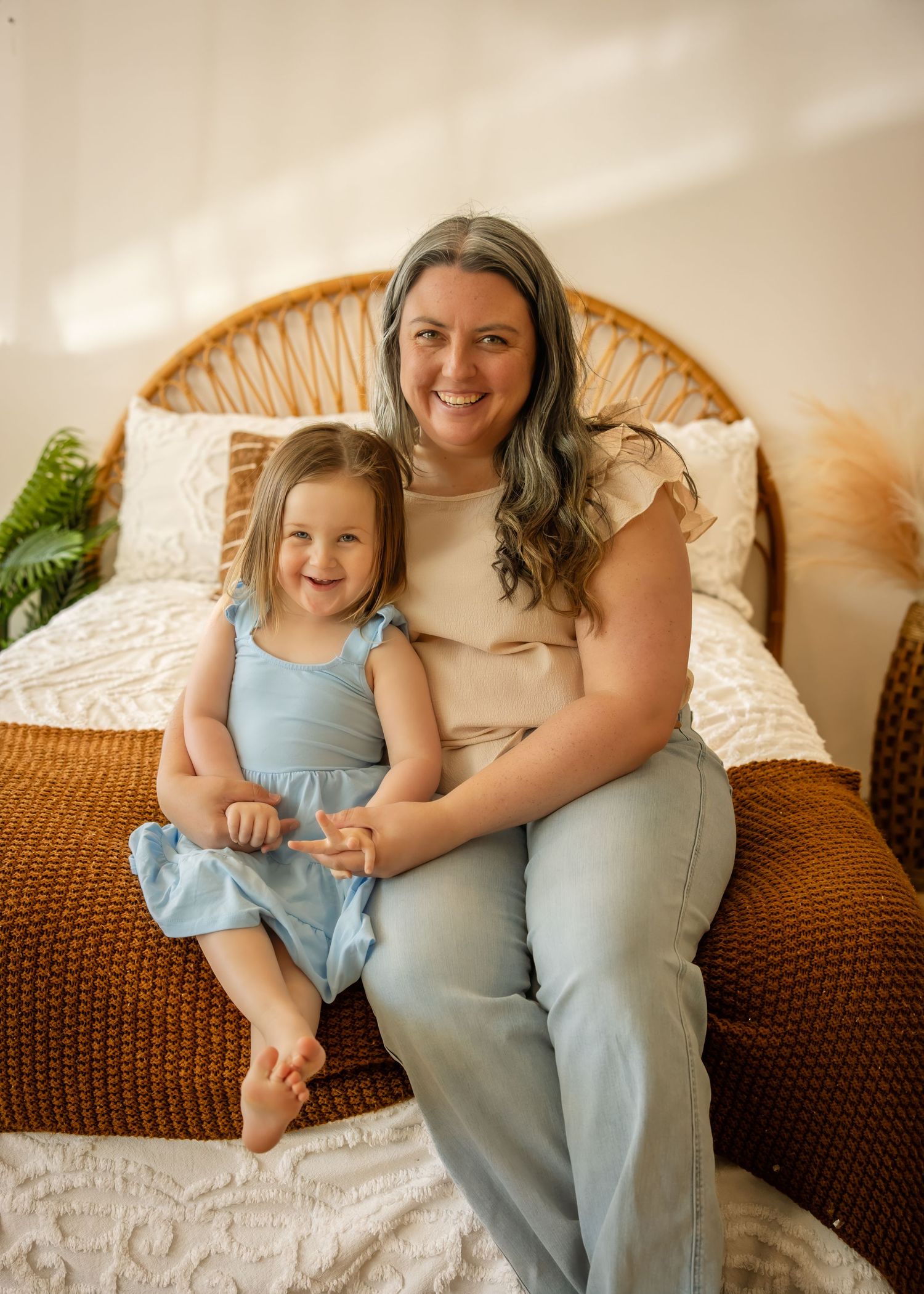A smiling woman and young girl in blue dress sit together on a bed with wicker headboard in a bright room.