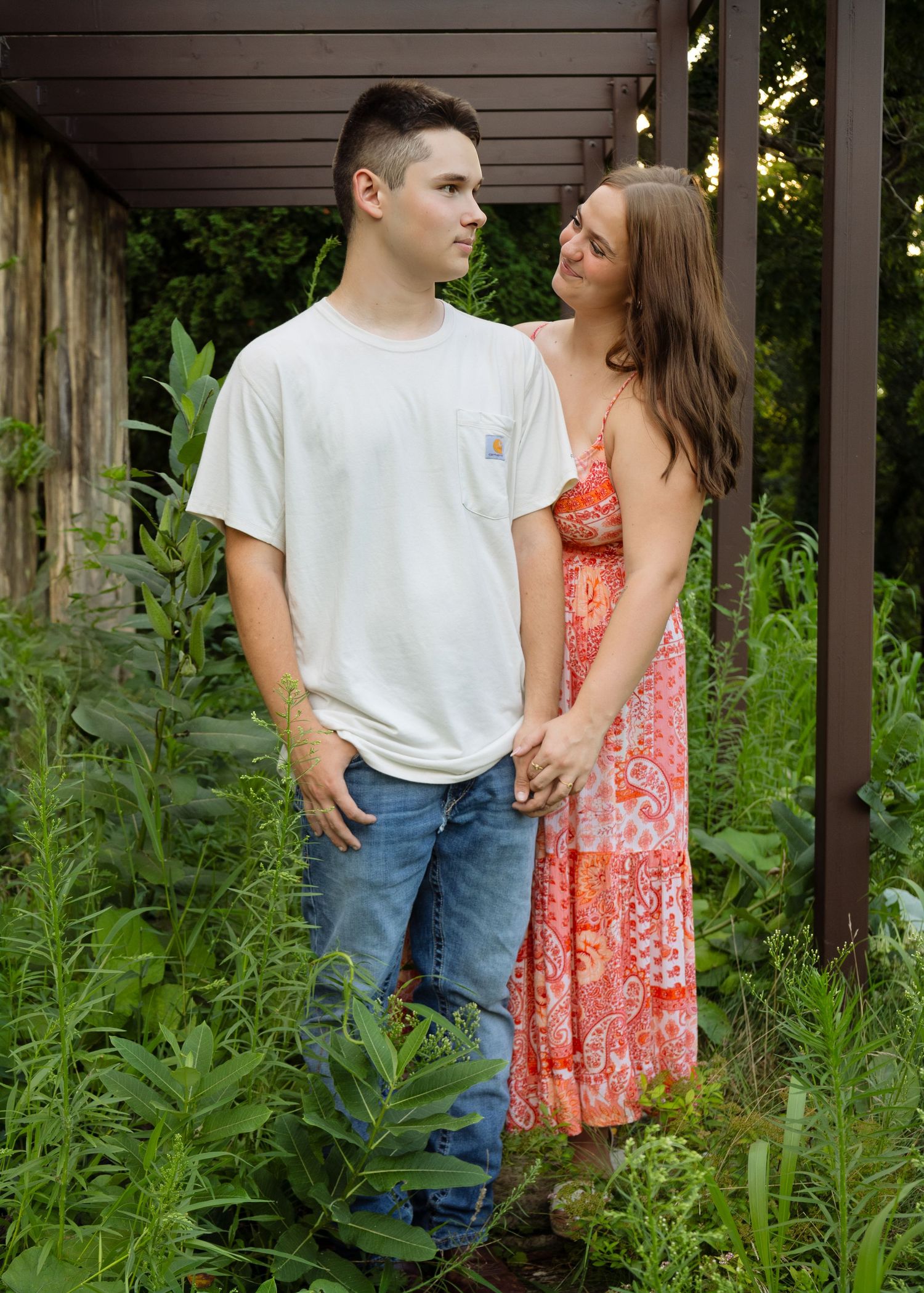 Young couple holding hands and gazing at each other in an overgrown garden beneath a pergola.