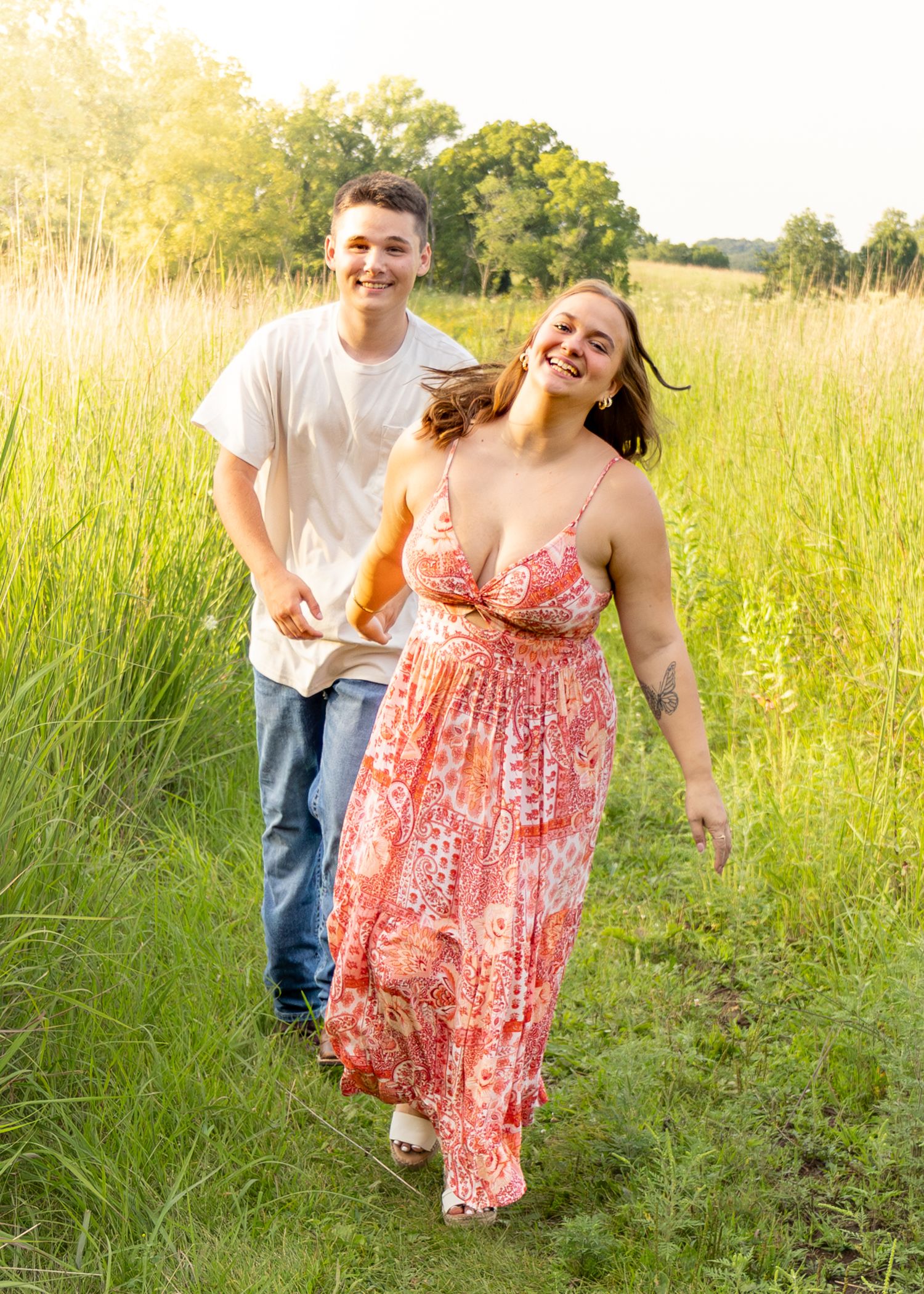 Happy couple walking hand in hand through a lush green meadow path, woman in red floral maxi dress.