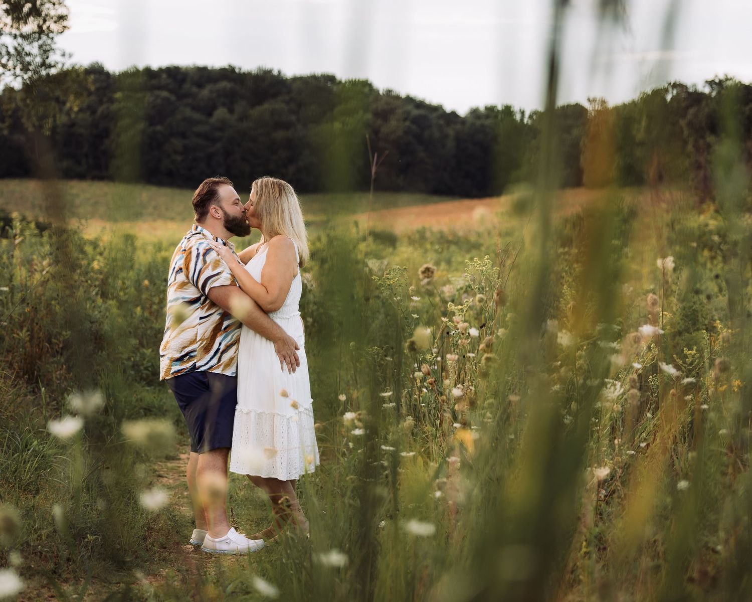 Couple embracing and kissing in a sunlit wildflower meadow surrounded by lush green trees.