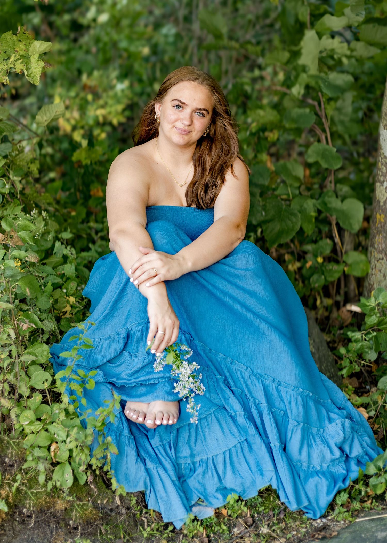 Young woman in a blue strapless gown holding white flowers, posed outdoors among lush green foliage.