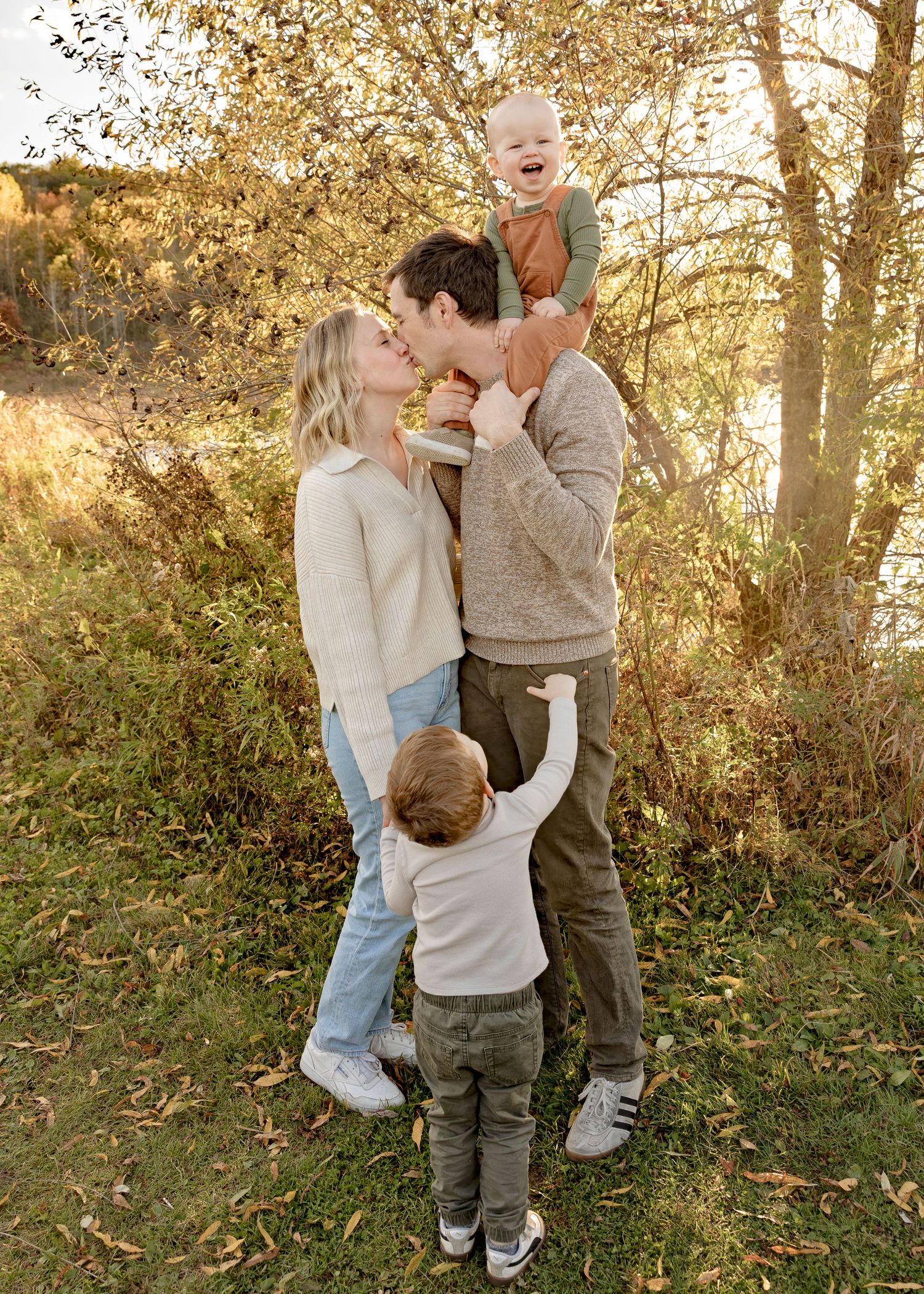 Family of four sharing a kiss outdoors in golden autumn light, toddler reaching up to parents.