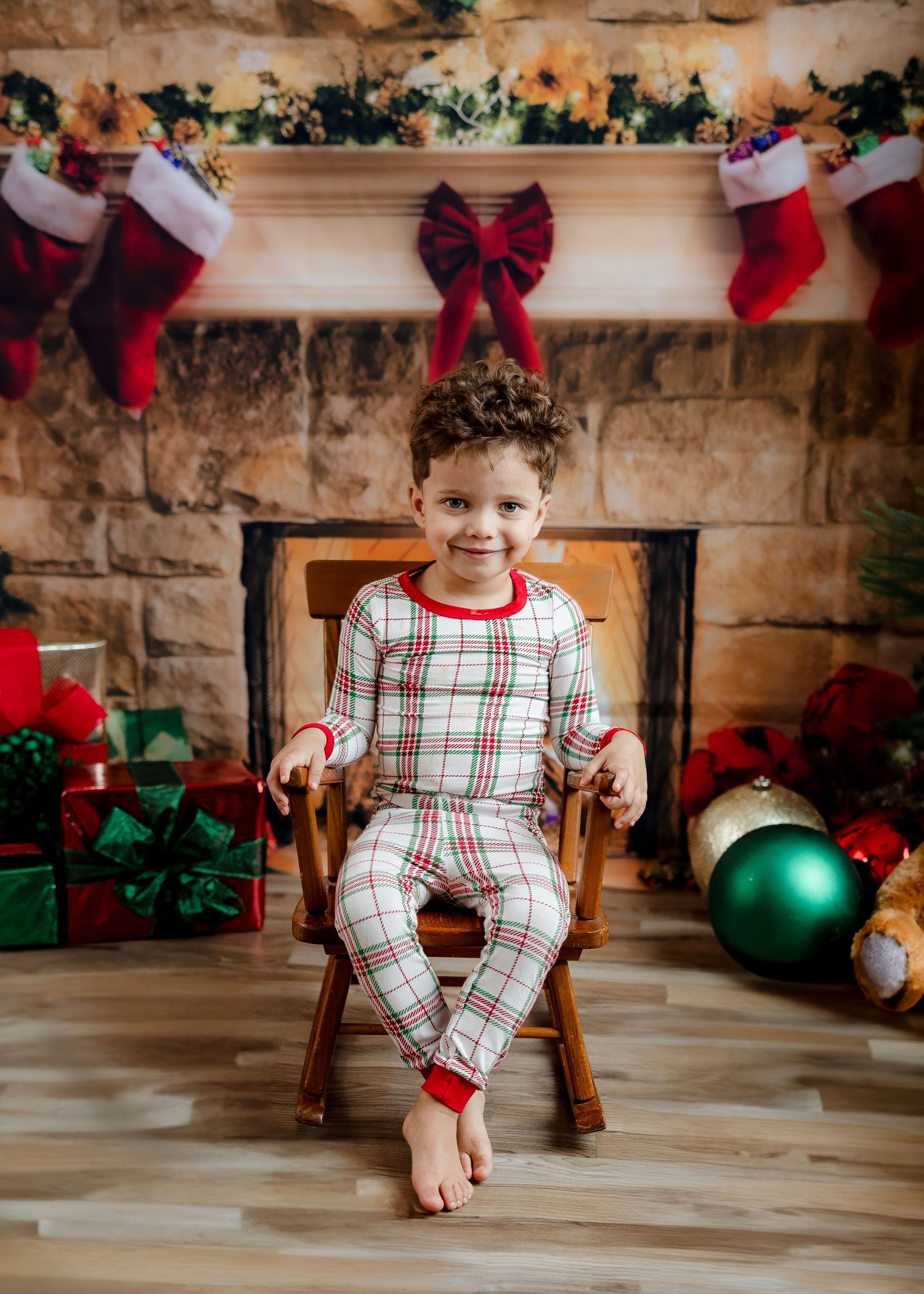 Young boy in plaid Christmas pajamas sitting barefoot in wooden chair by festive fireplace.