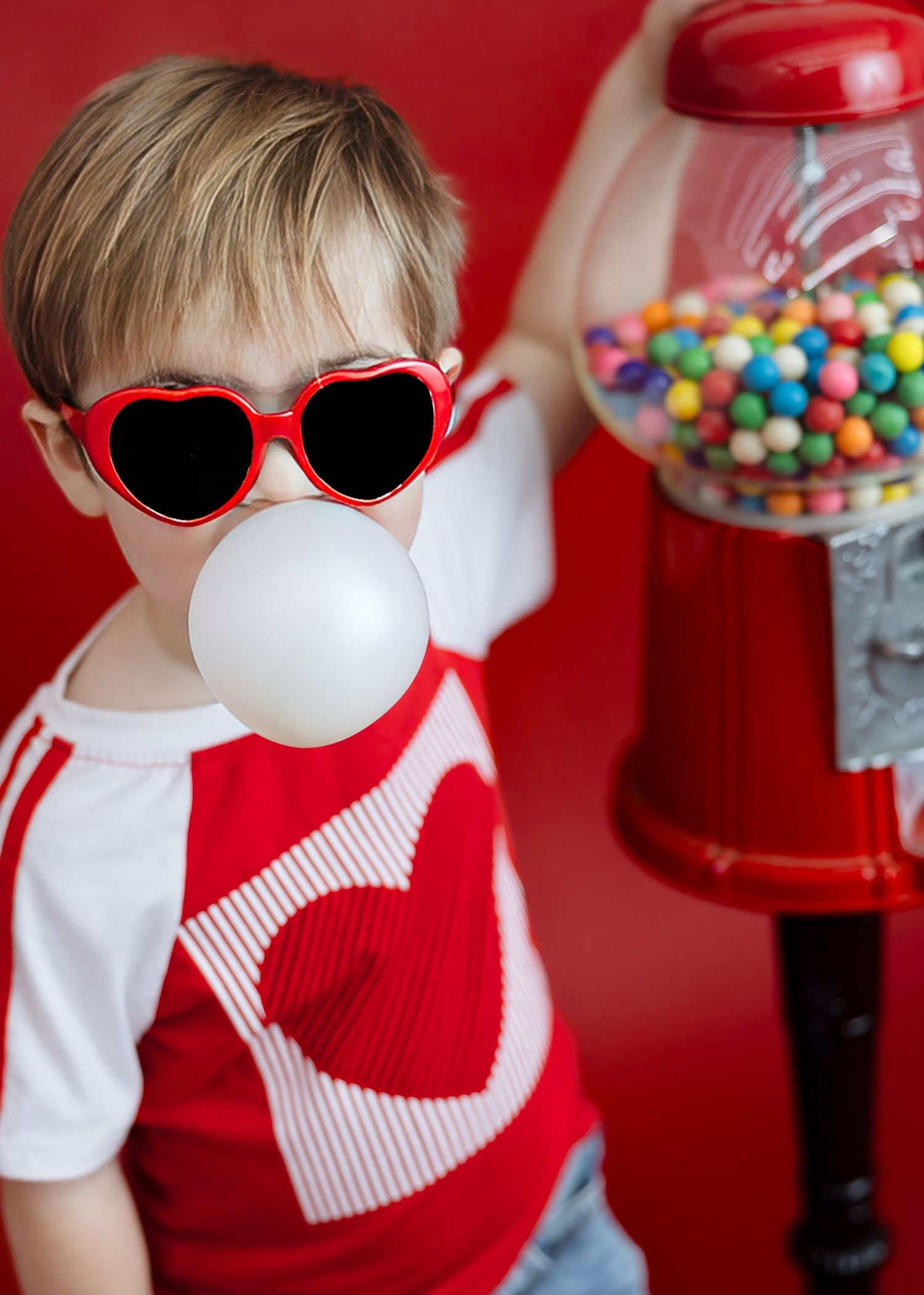 Trendy boy in heart sunglasses blowing bubble gum bubble next to colorful gumball machine.