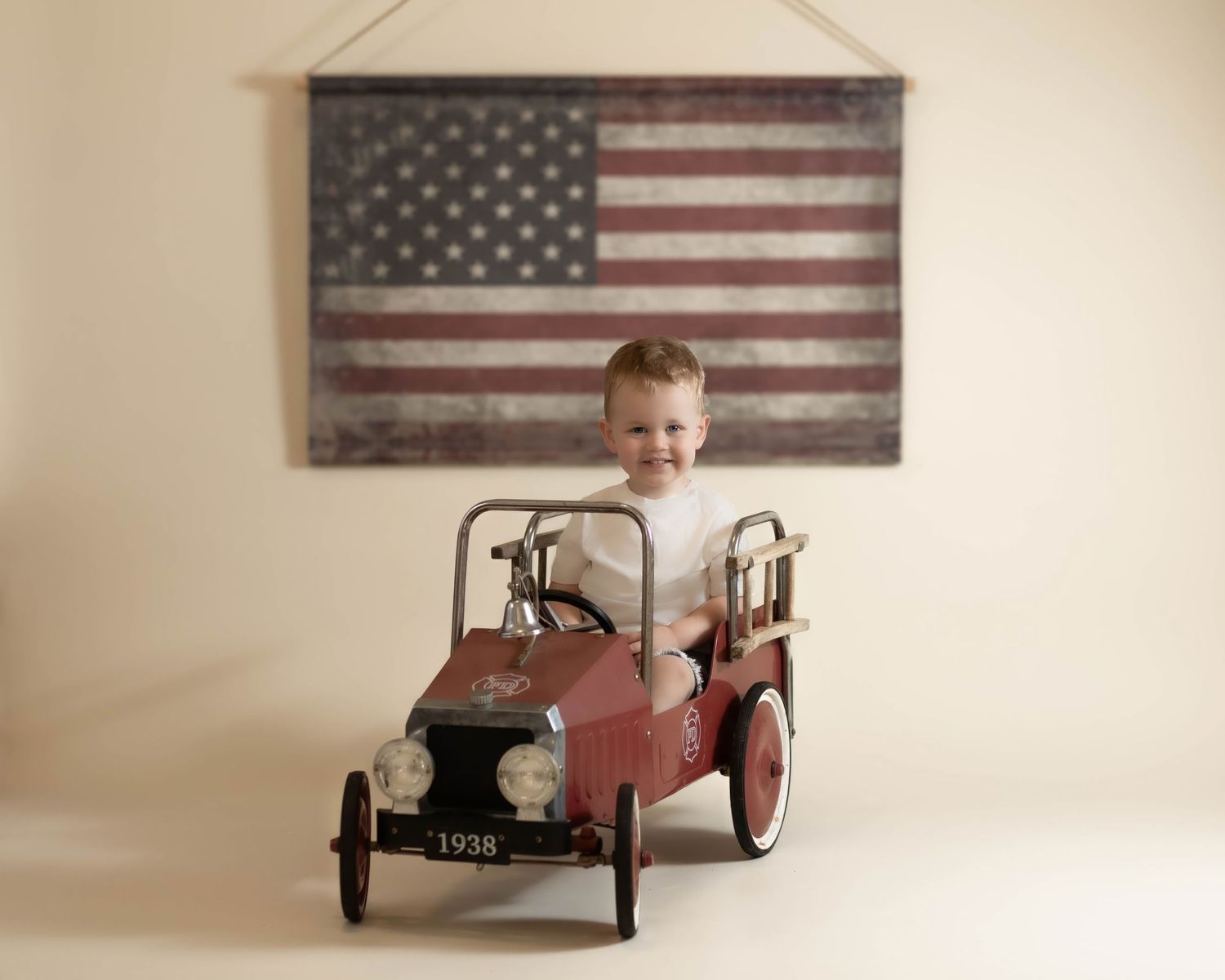 Smiling toddler boy in white shirt rides a vintage red pedal fire truck before an American flag.