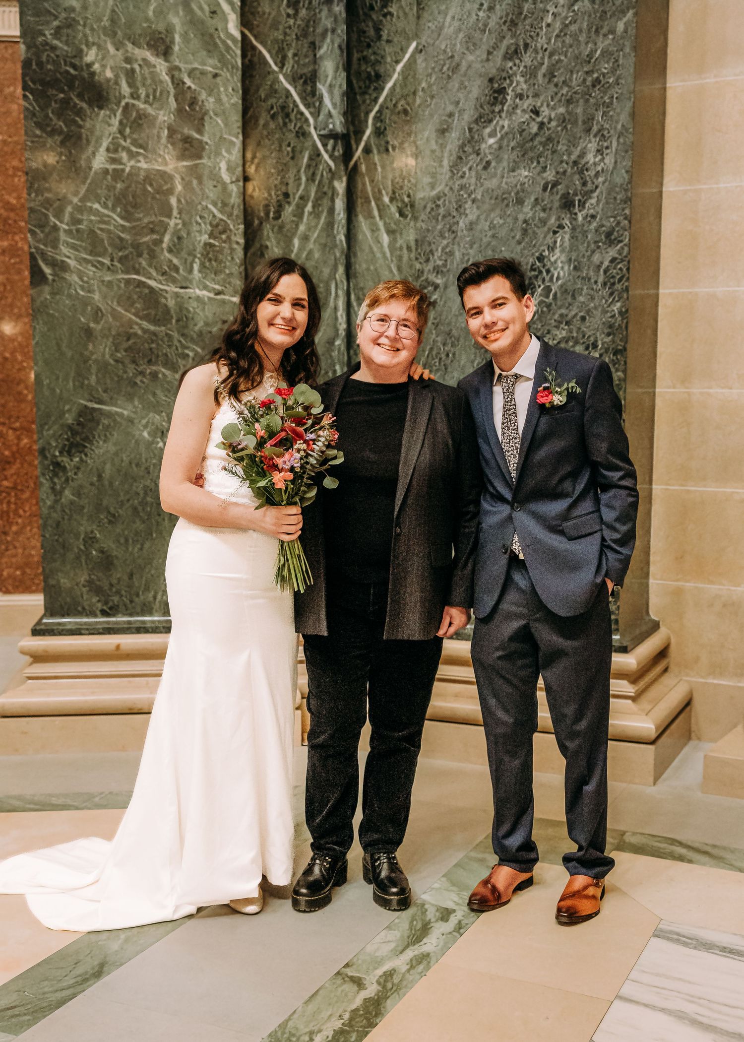 Bride with bouquet, officiant, and groom pose together smiling before marble column backdrop.