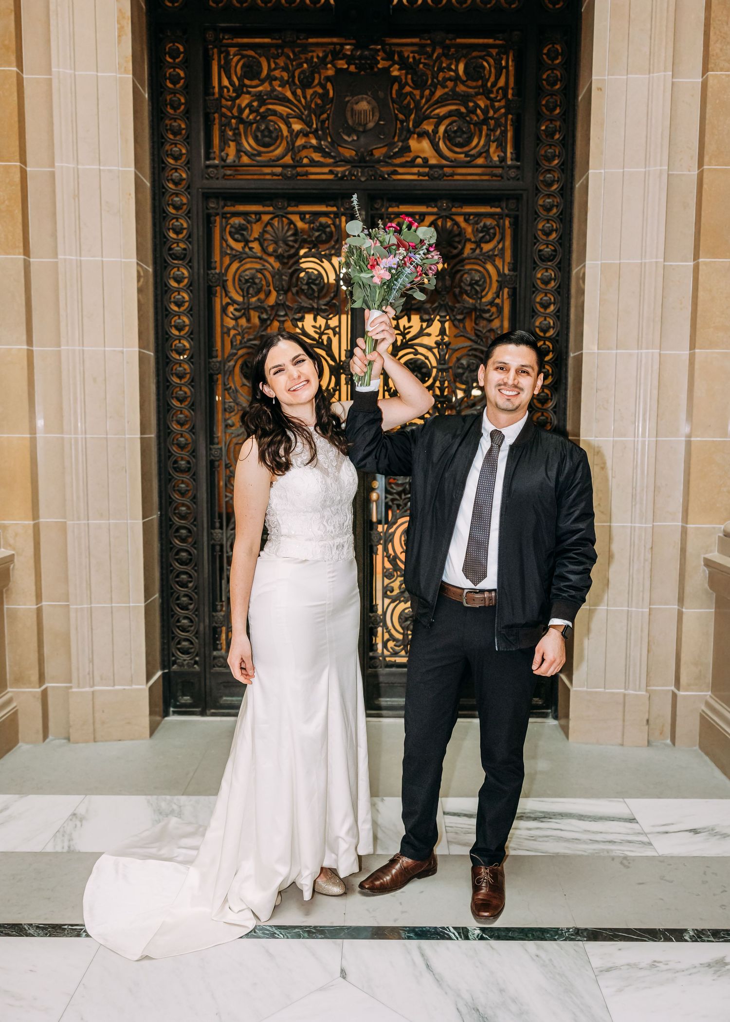 Smiling bride and groom celebrating with raised floral bouquet before ornate golden doors.