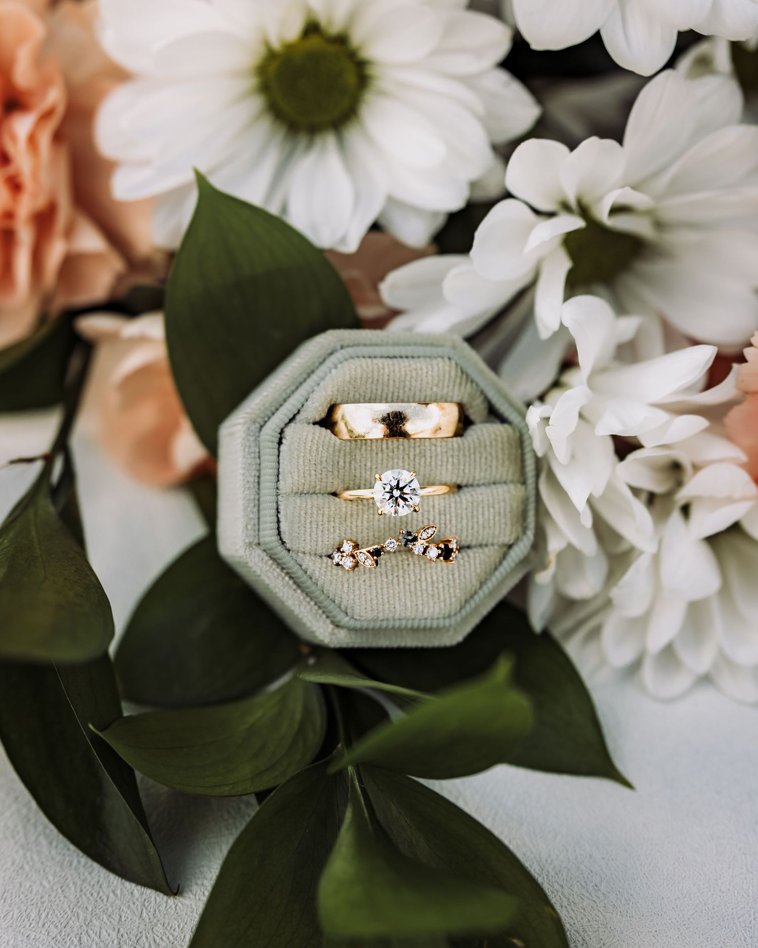 Three gold rings displayed in a sage green velvet octagonal box surrounded by white daisies and peach roses.
