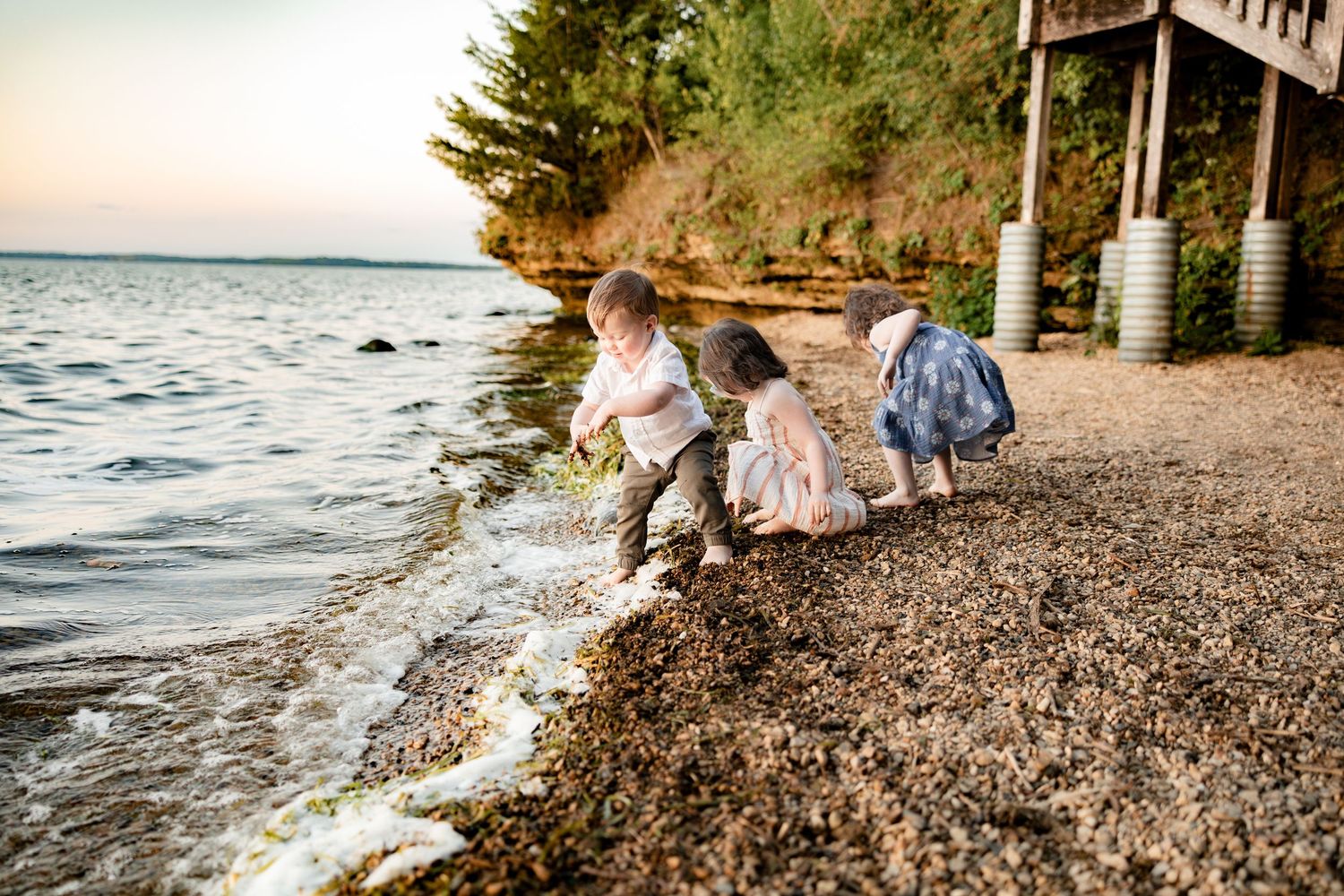 Three young children playing at the rocky shoreline, crouching by gentle waves at sunset near a wooden dock.