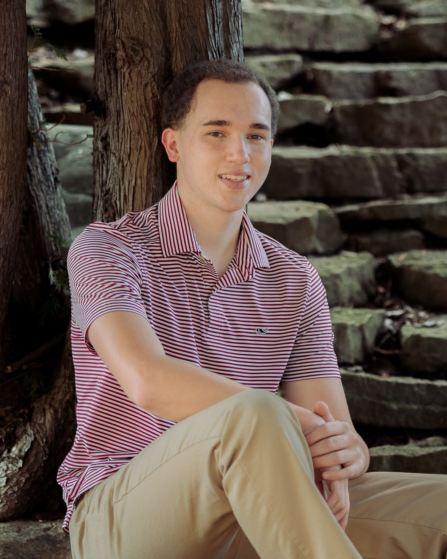 Young man in red striped polo shirt sitting outdoors against a tree with stone steps in background.