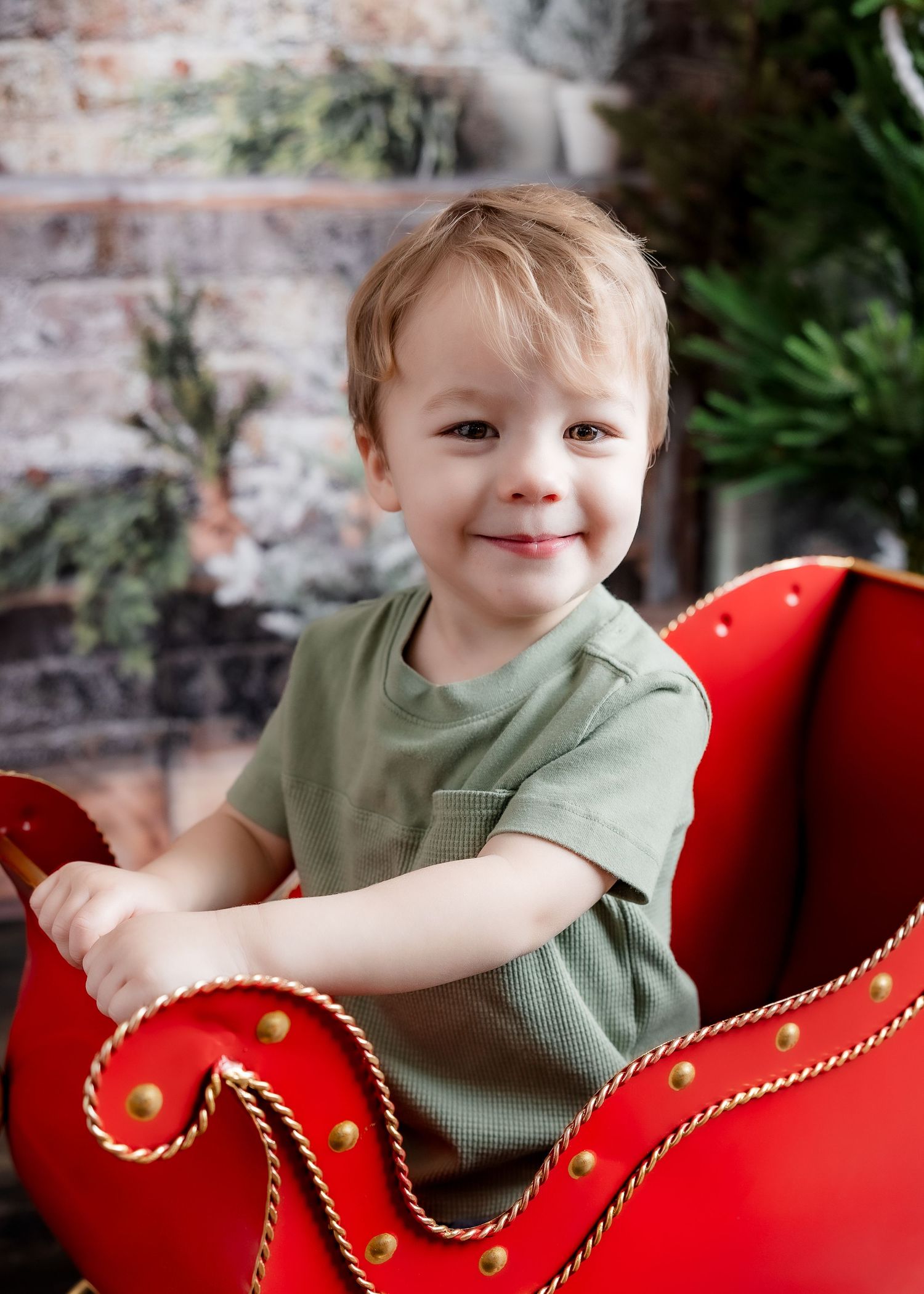 A cute smiling toddler boy sits in a decorative red and gold Christmas sleigh against a brick wall backdrop.