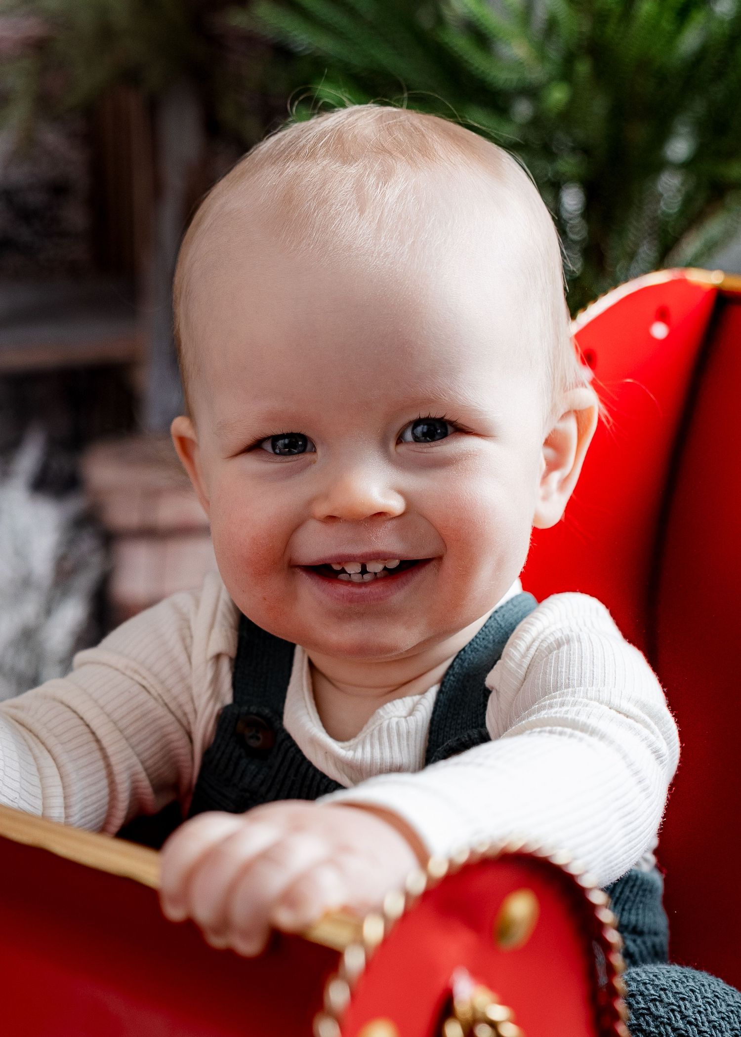 Smiling baby boy with two teeth sitting in a red Christmas sleigh wearing green overalls.