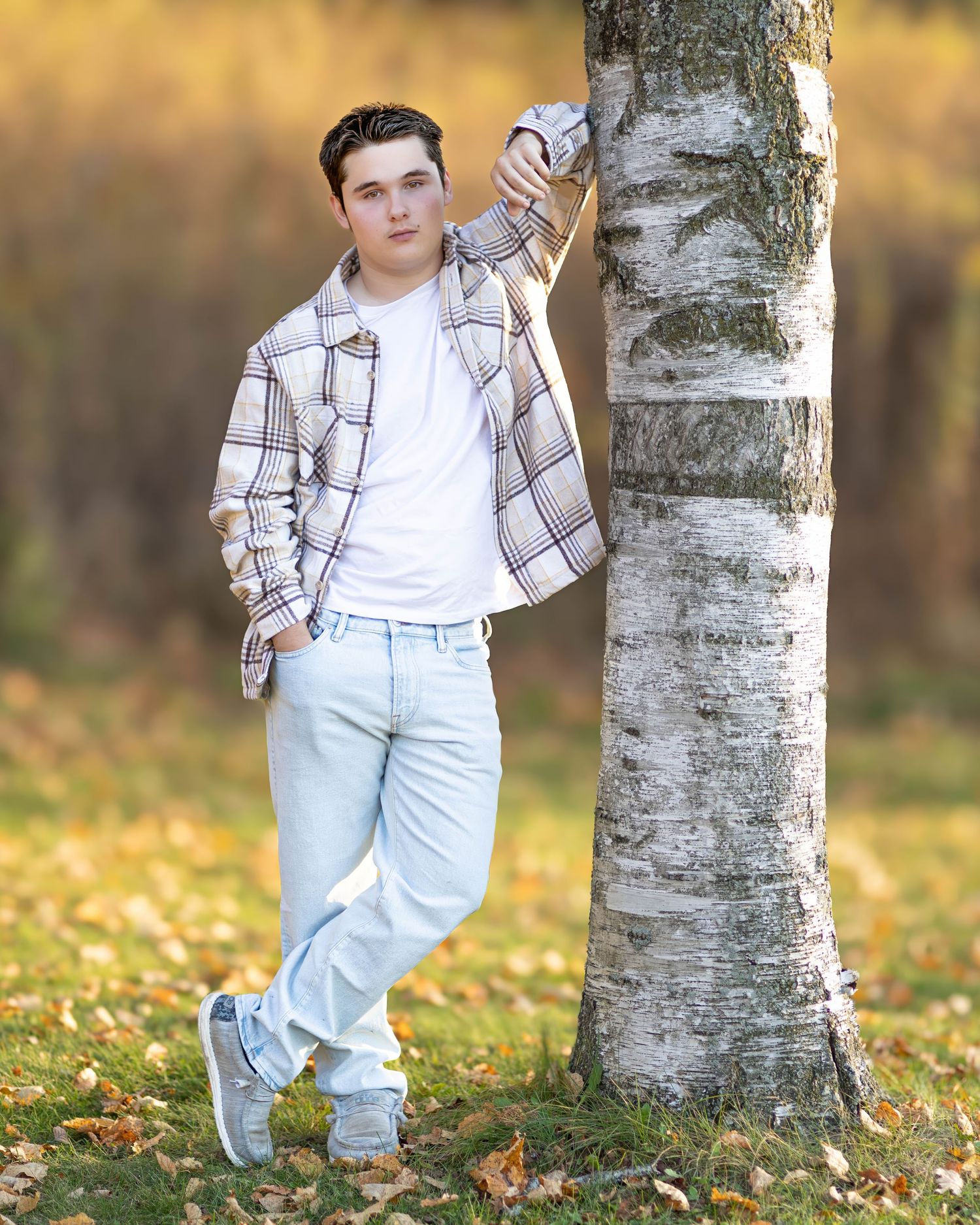 Teen boy in plaid shirt leans against birch tree surrounded by autumn leaves.