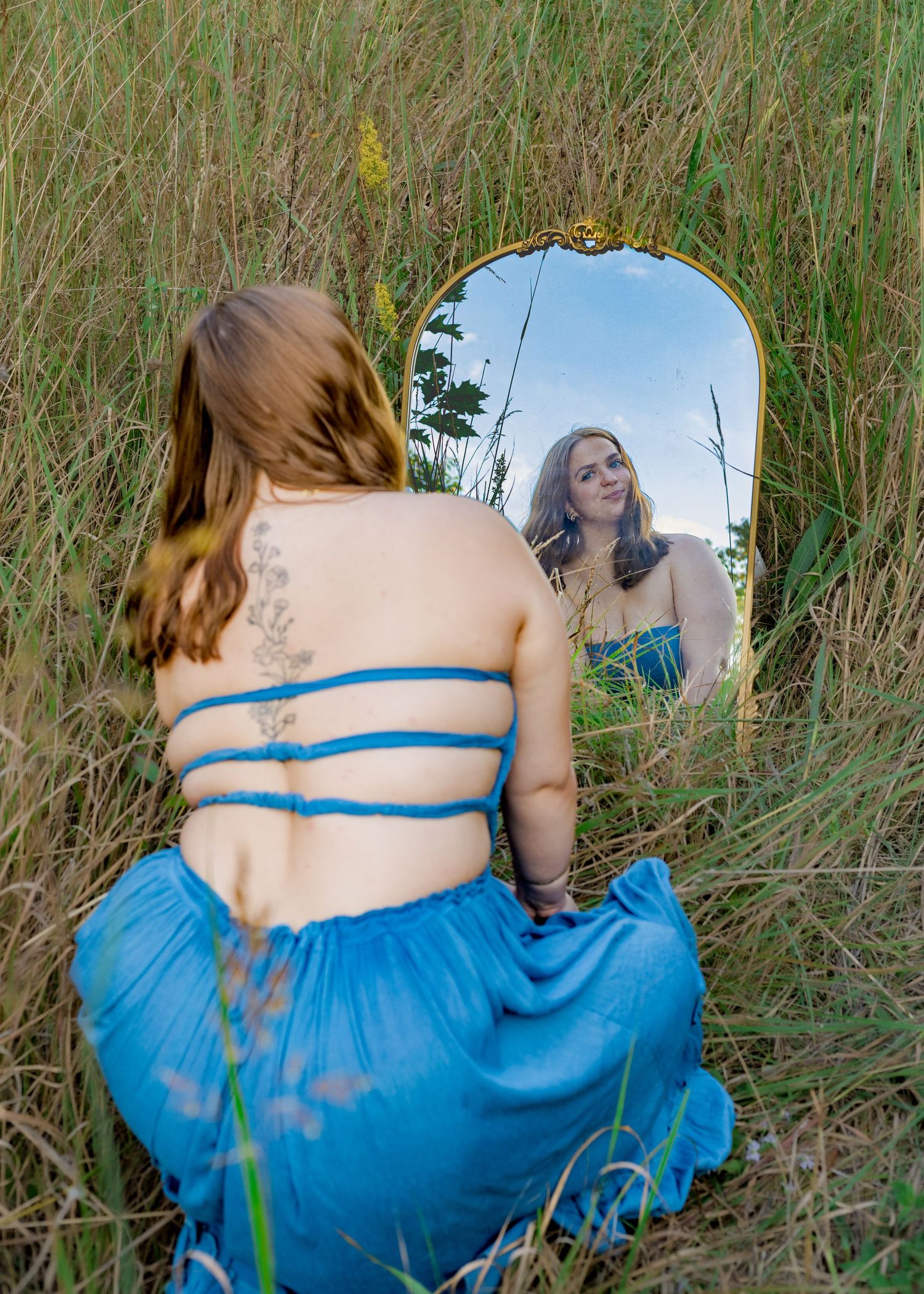 Senior girl in blue dress sits in tall grass viewing her reflection in ornate gold mirror.