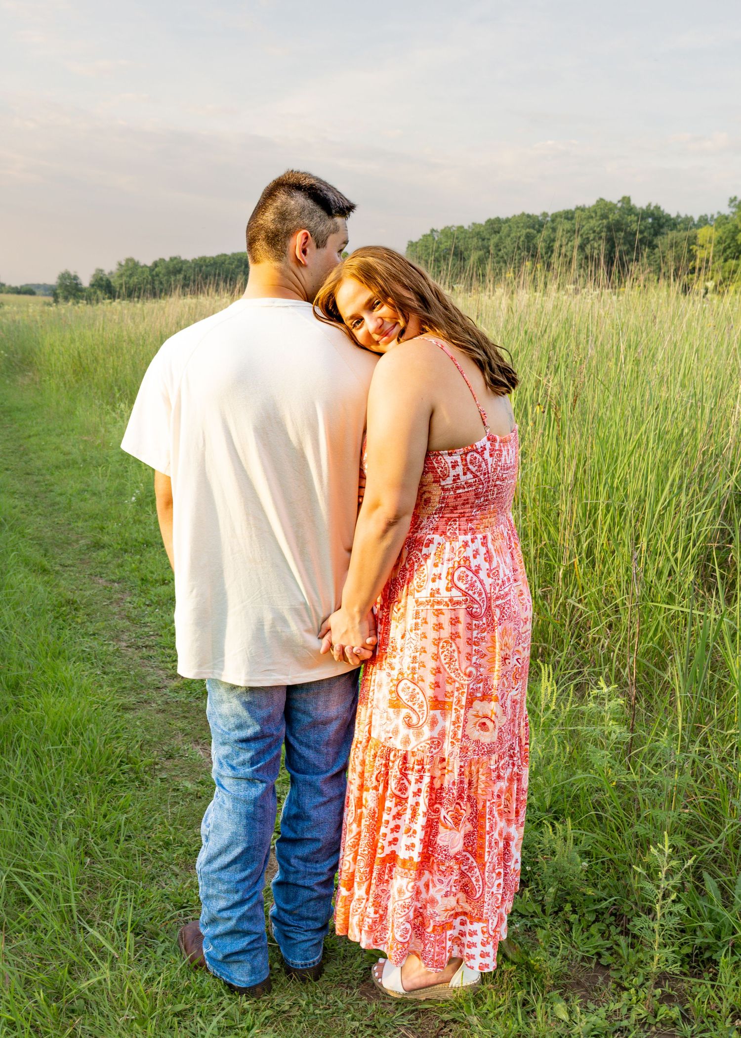 Couple holding hands on a grassy field path, woman in pink maxi dress leaning on mans shoulder at golden hour.
