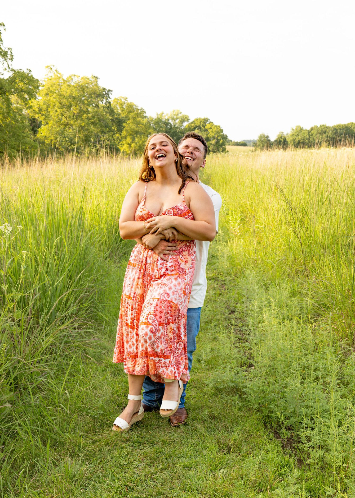Smiling couple hugging on a grassy field path at sunset, woman in red floral dress leaning back.