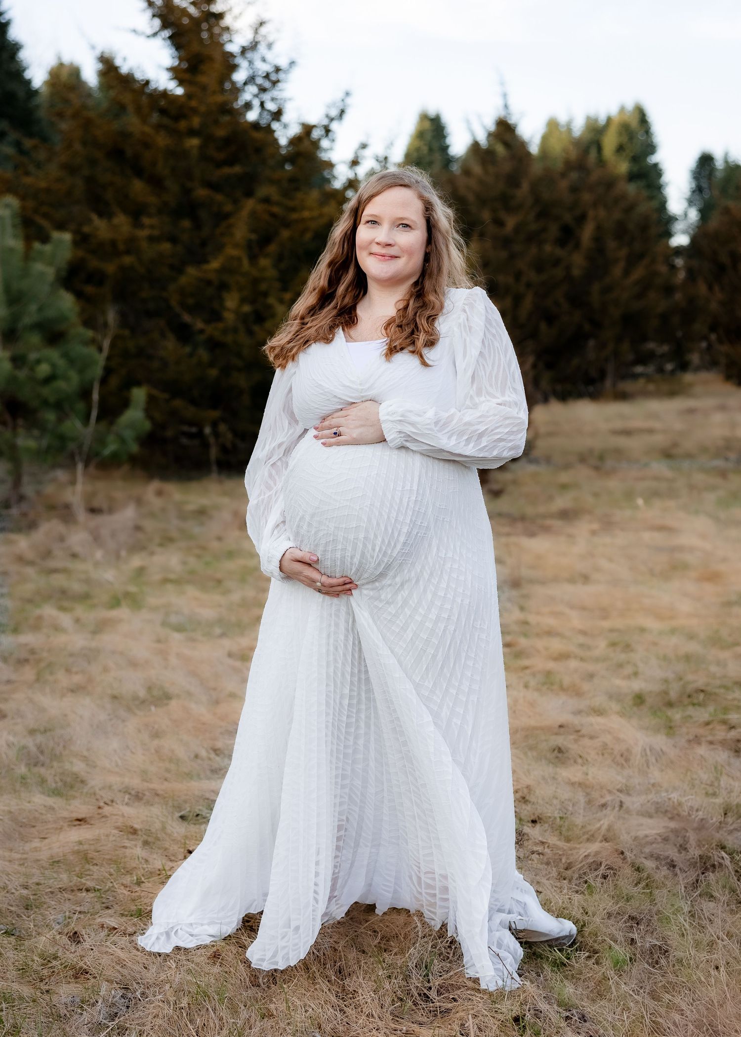 Pregnant woman in white lace maternity gown standing in field with evergreen trees behind her.